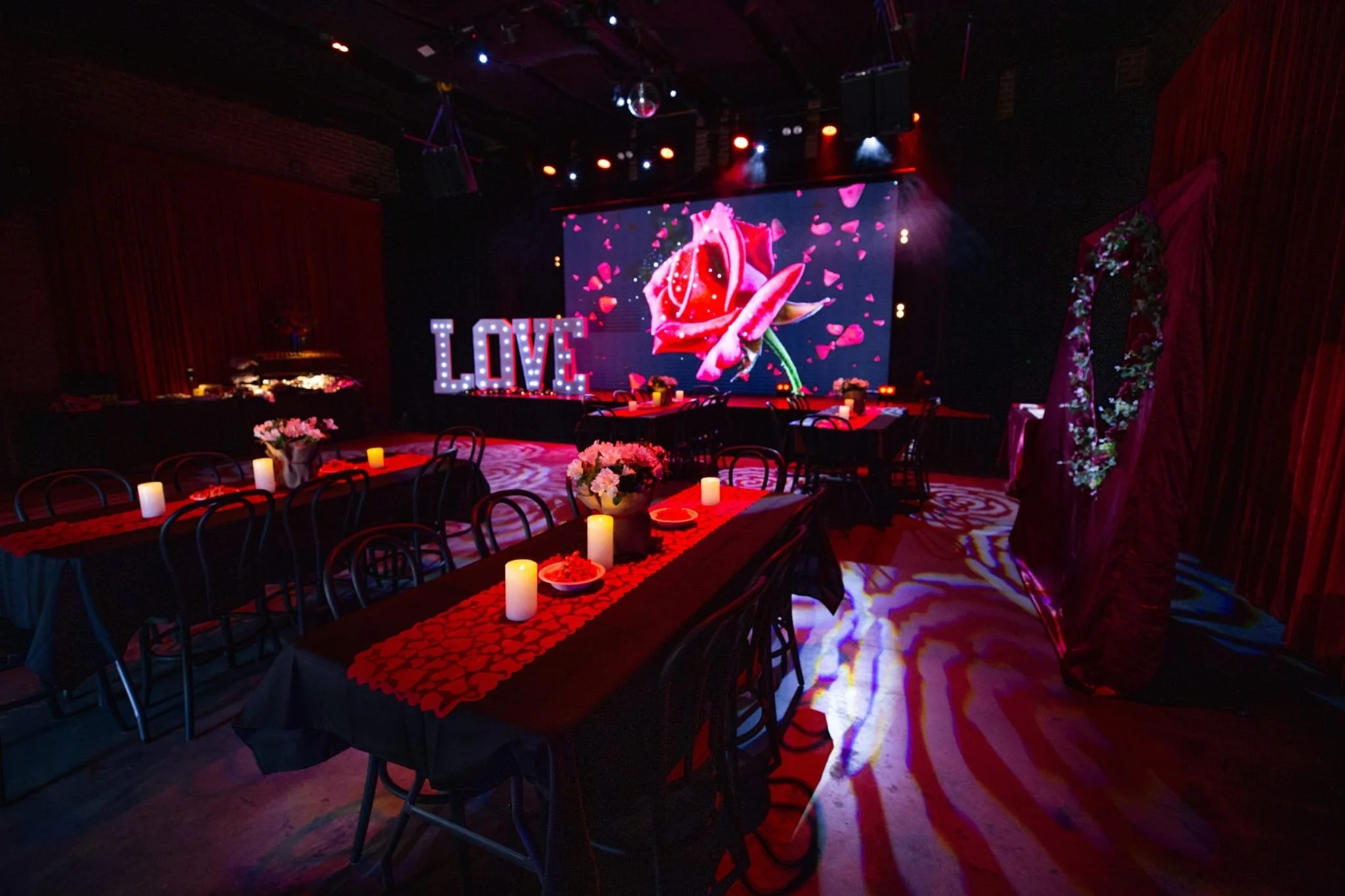 A dimly lit event venue decorated for a romantic occasion, featuring long tables with black tablecloths, red heart runners, and candles. A stage in the background displays large light-up "LOVE" letters and a screen showing a red rose, while the floor