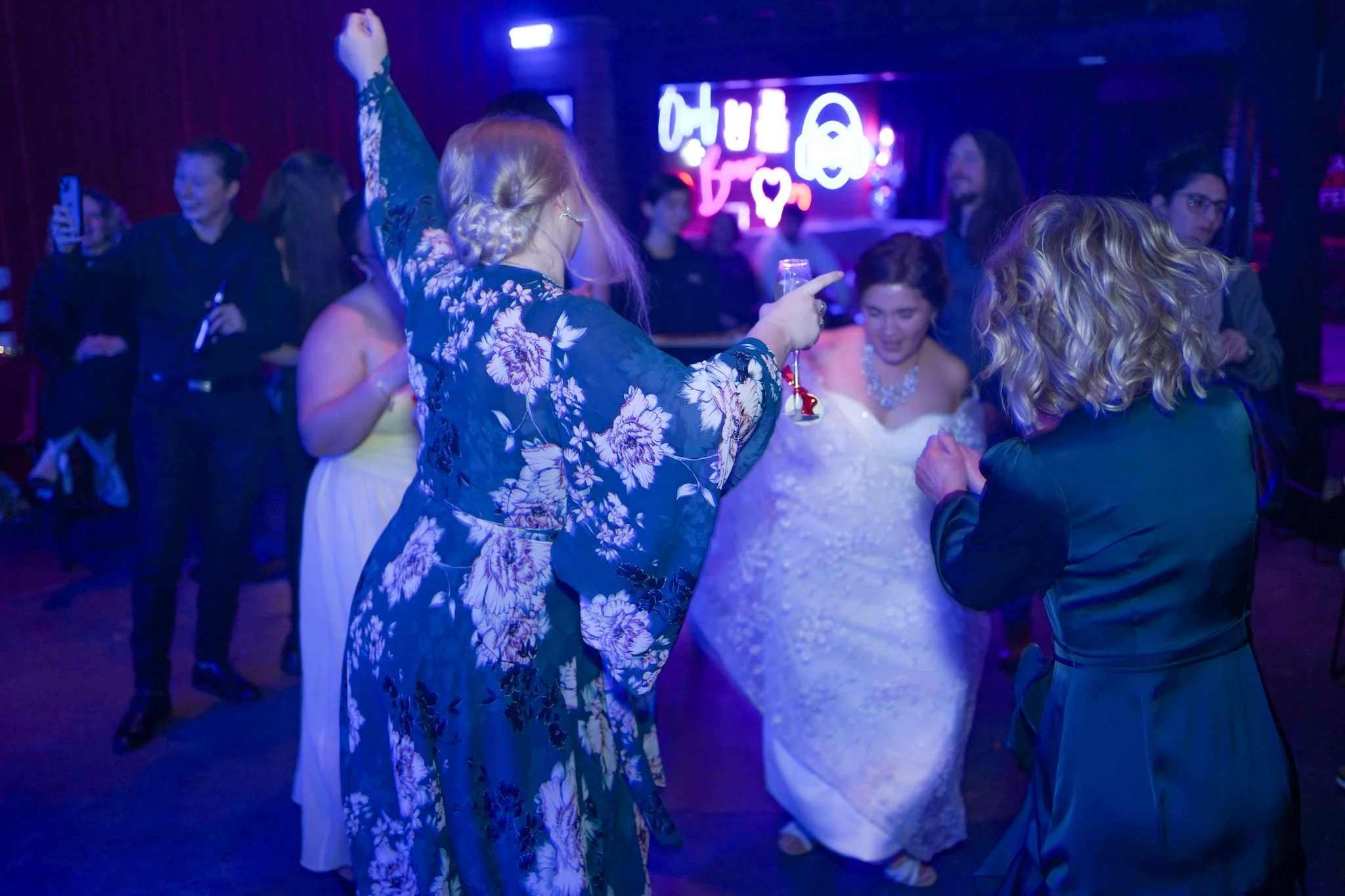 Bride and friends dancing together under moody blue lighting in front of vibrant neon signage.