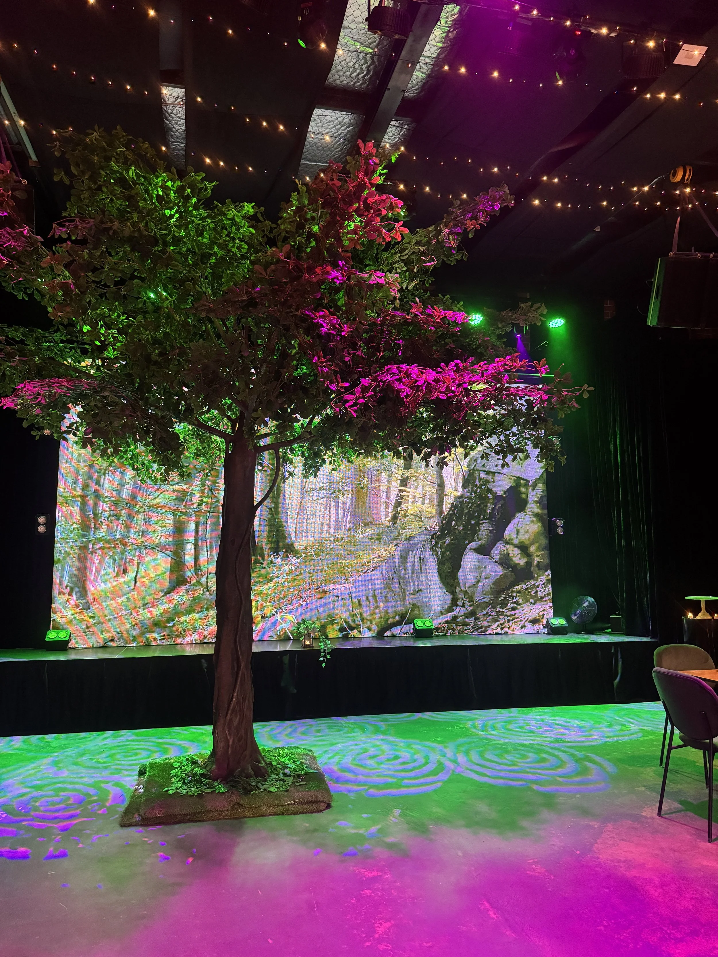 A leafy artificial tree illuminated by pink and green spotlights stands on a green patterned light projection. Behind the tree, a large LED stage screen displays a detailed forest landscape under warm venue ceiling lights.