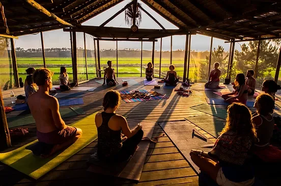 Menschen üben Yoga in einer offenen, hölzernen Struktur bei Sonnenaufgang im Freien, mit Blick auf grüne Felder