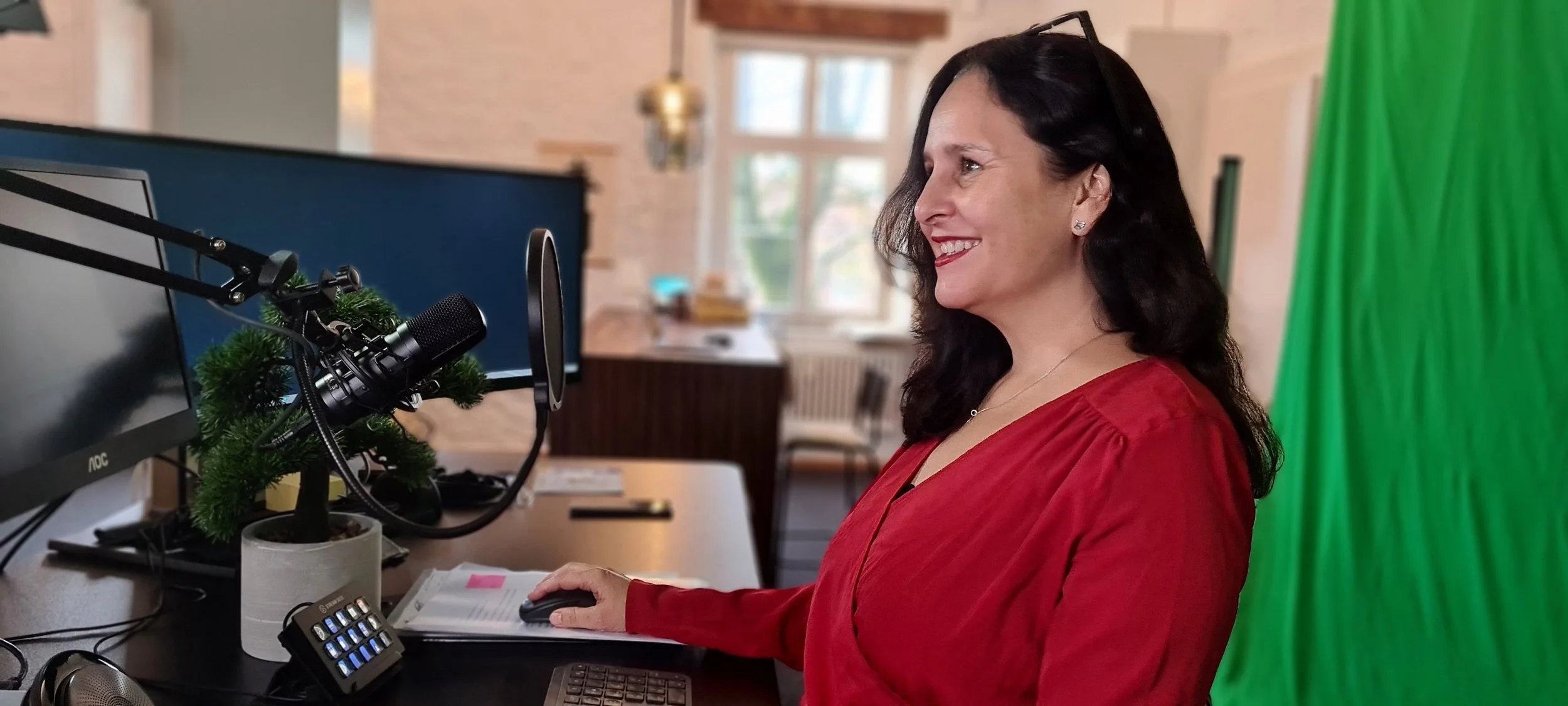 Eine Frau sitzt vor einem Computerbildschirm in einem Raum mit einem grünen Bildschirm im Hintergrund, vermutlich für einen Videoaufnahme- oder Streaming-Aufnahme.