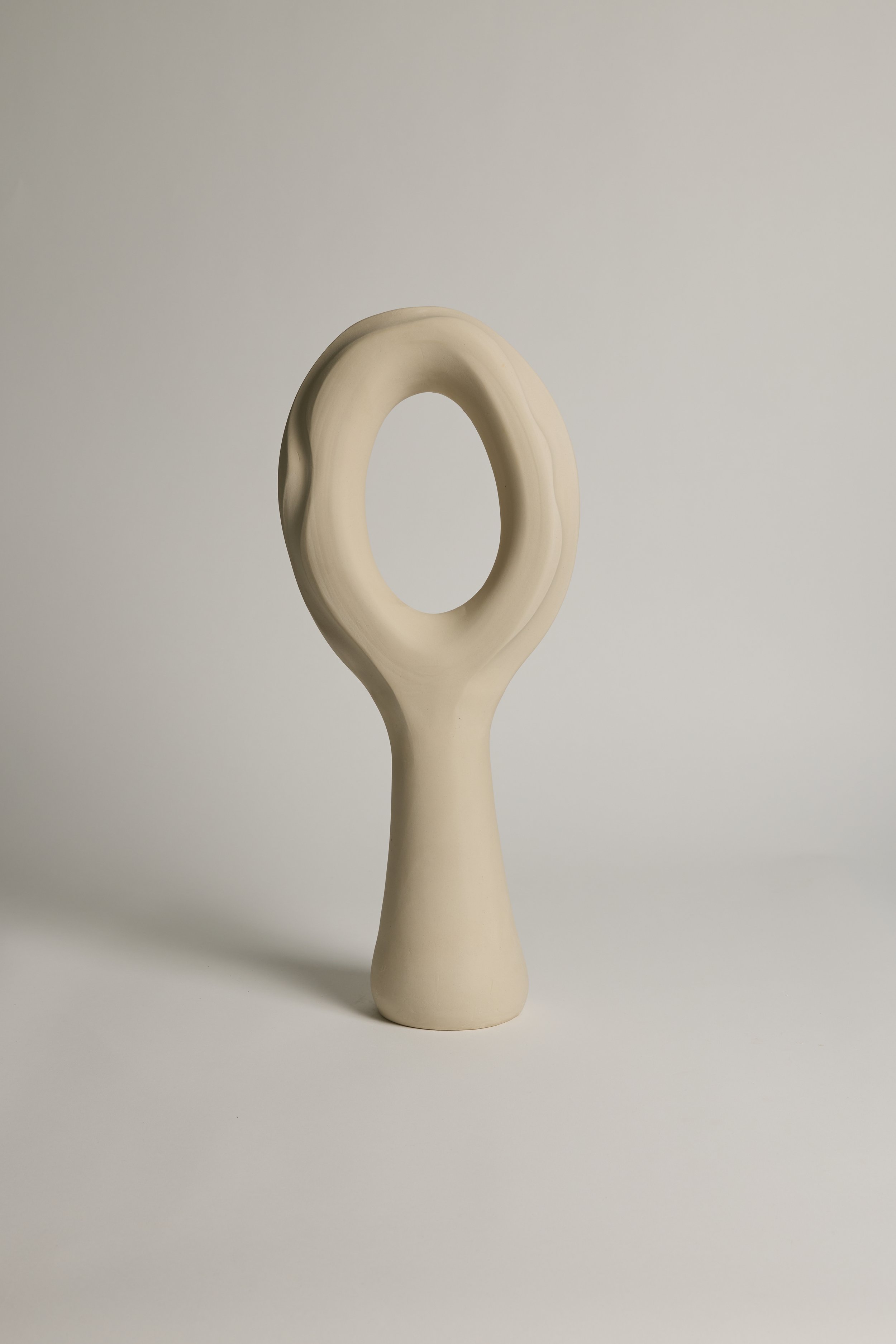 A cream-colored, abstract ceramic sculpture with a looped design, standing on a flat base against a plain light background.