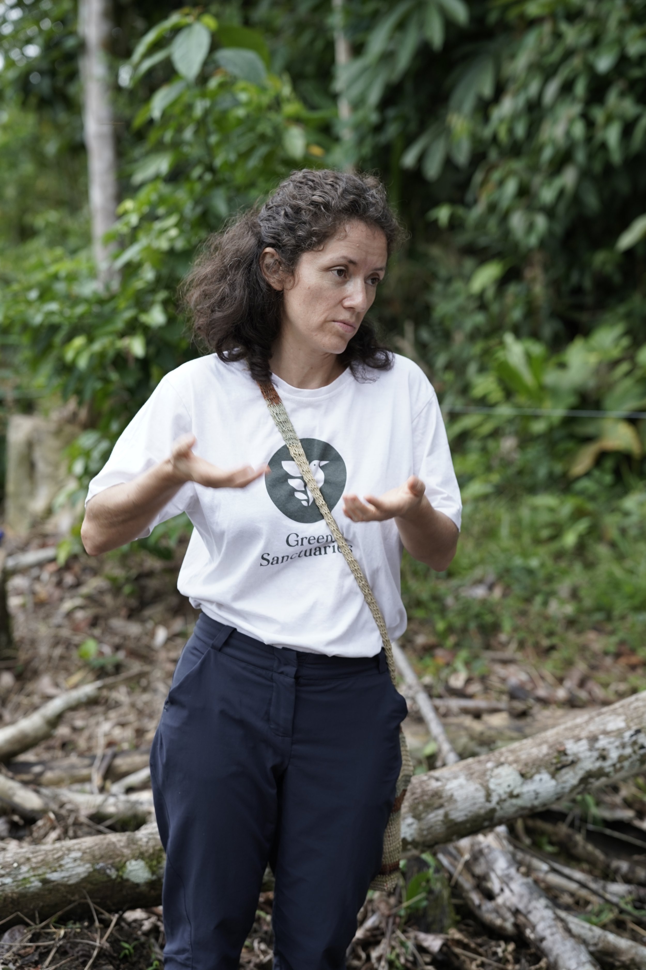 Femme souriante portant un t-shirt blanc avec un logo d'oiseau et la mention 'Green Sanctuaries', entourée de plusieurs personnes dans un cadre extérieur.