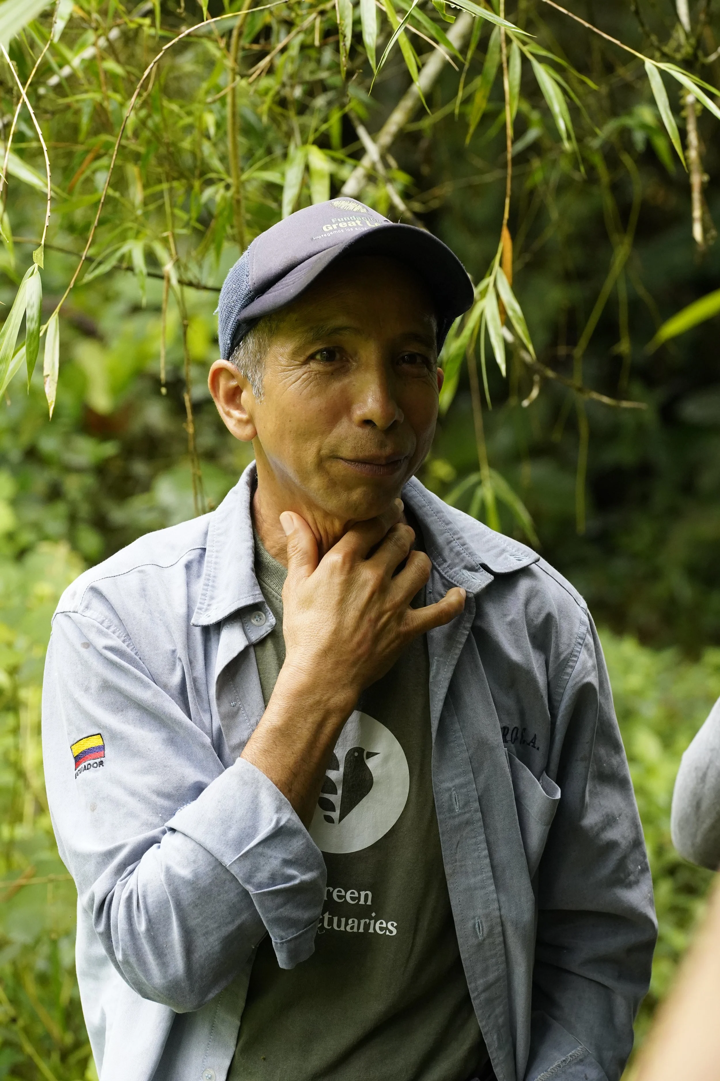 Un homme portant un t-shirt blanc et une casquette blanche, faisant un geste de paix avec la main, assis dans un environnement naturel avec des plantes en arrière-plan.