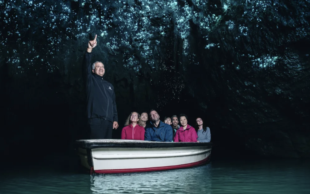 A group of people in a boat inside a dark cave with glowing blue water and bioluminescent cave formations, accompanied by a guide pointing upward.