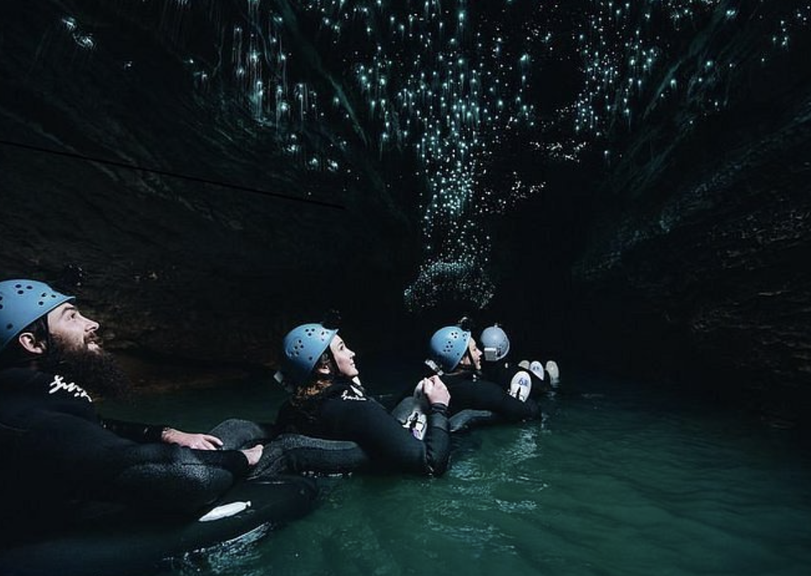 Group of five people in wetsuits and helmets lying on their stomachs on floatation devices, looking upward inside a dark cave with glowing blue water reflections on the ceiling.