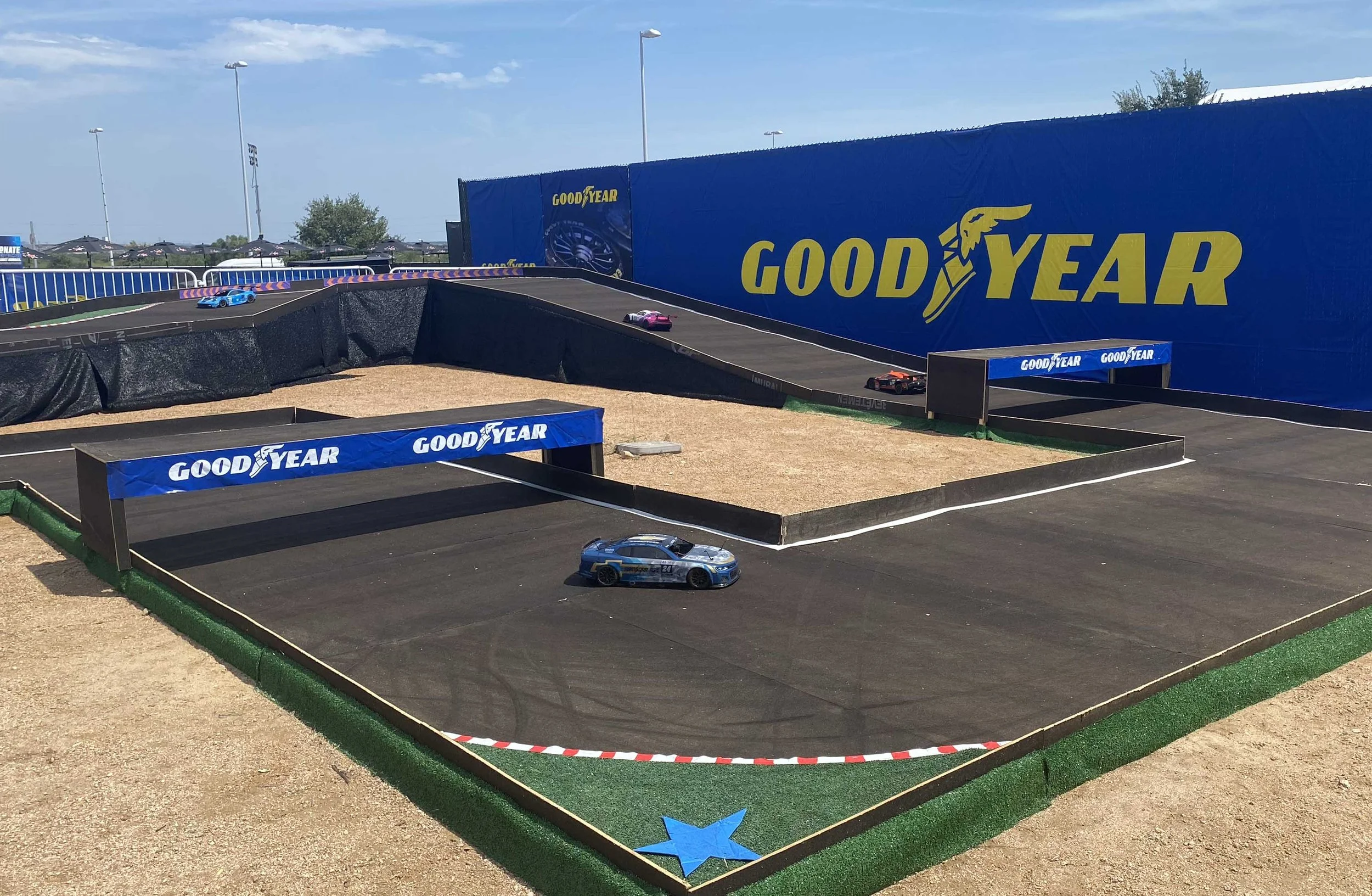 Young boy in racing seat playing a car racing game on a screen, with a green remote control car on a track in front of him, and two people standing nearby.