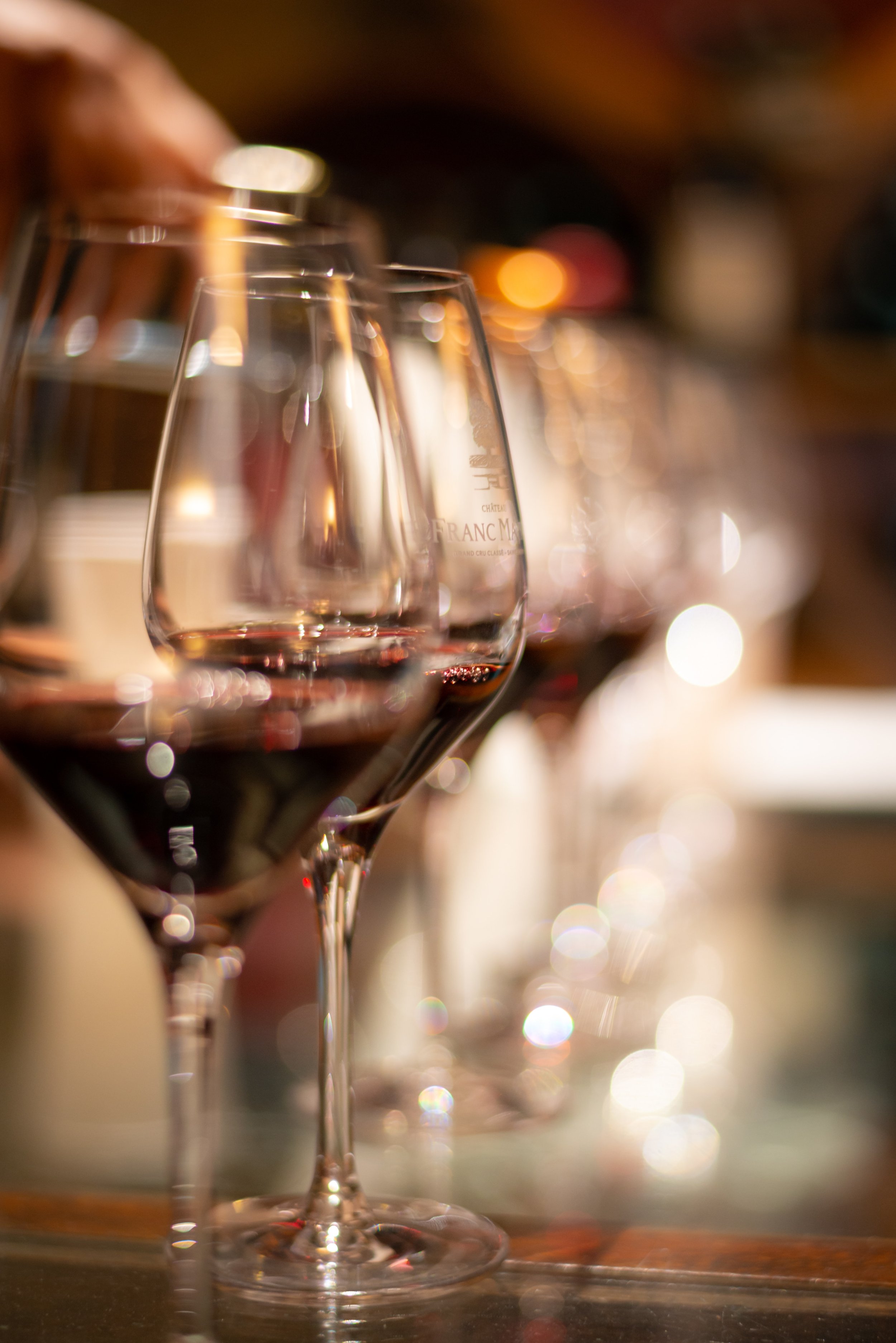 Row of wine glasses filled with red wine on a wooden surface, with a blurred background of a wine cellar or bar.