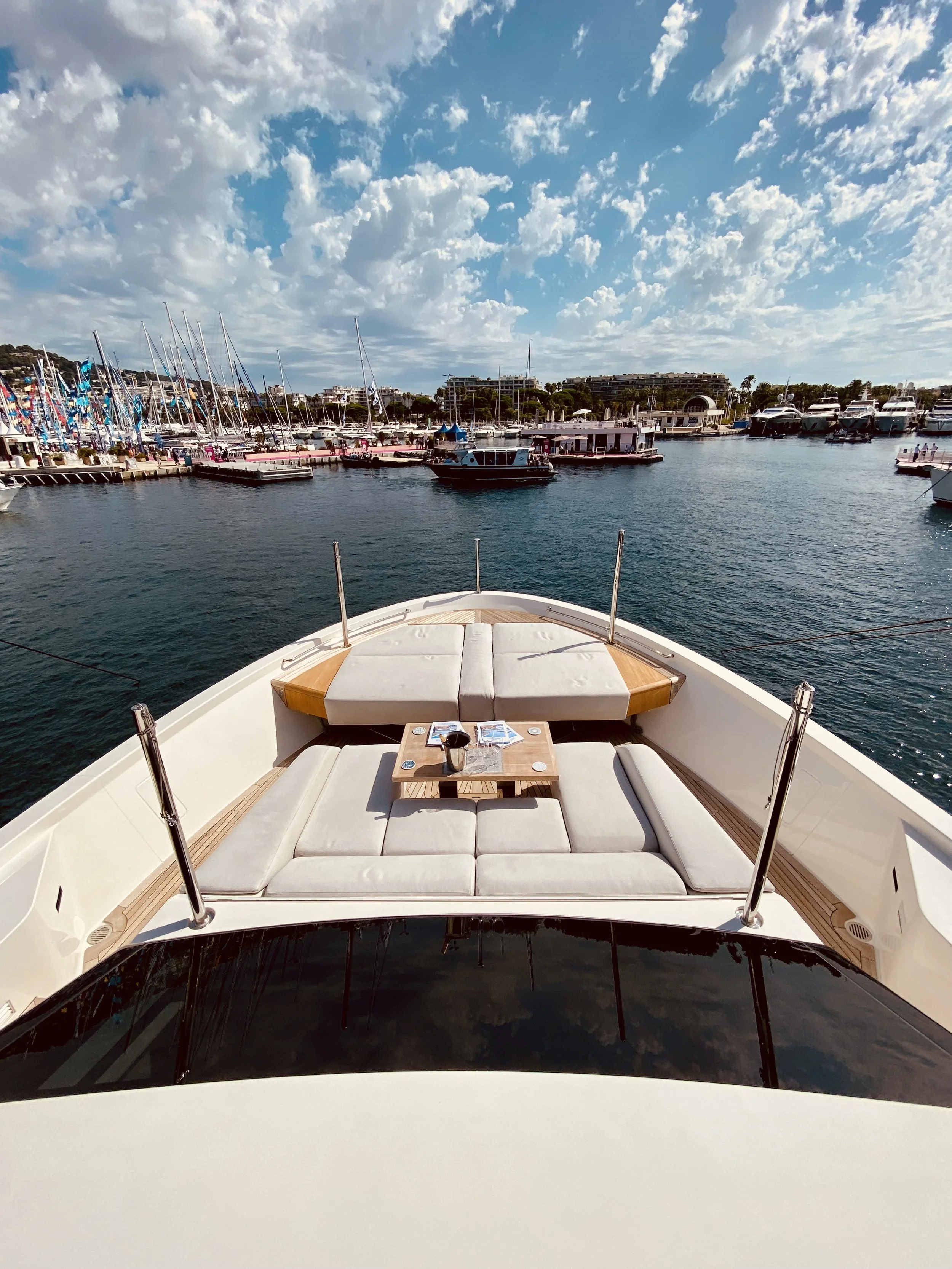 View from the bow of a yacht with outdoor seating and a small table, overlooking a marina filled with sailboats and yachts under a partly cloudy sky.
