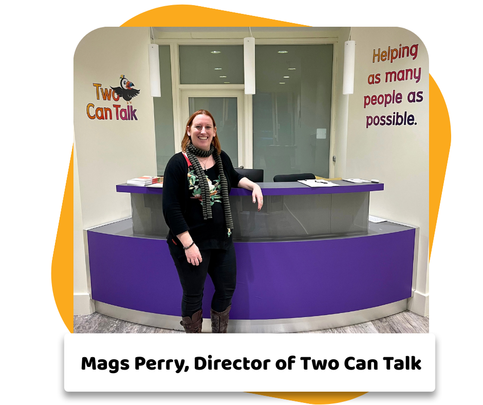 Mags Perry standing at the reception desk of Two Can Talk resource center, smiling, behind a purple counter. Wall decorations include a colorful sign reading "Two Can Talk" with a bird logo and a motivational quote about helping people.