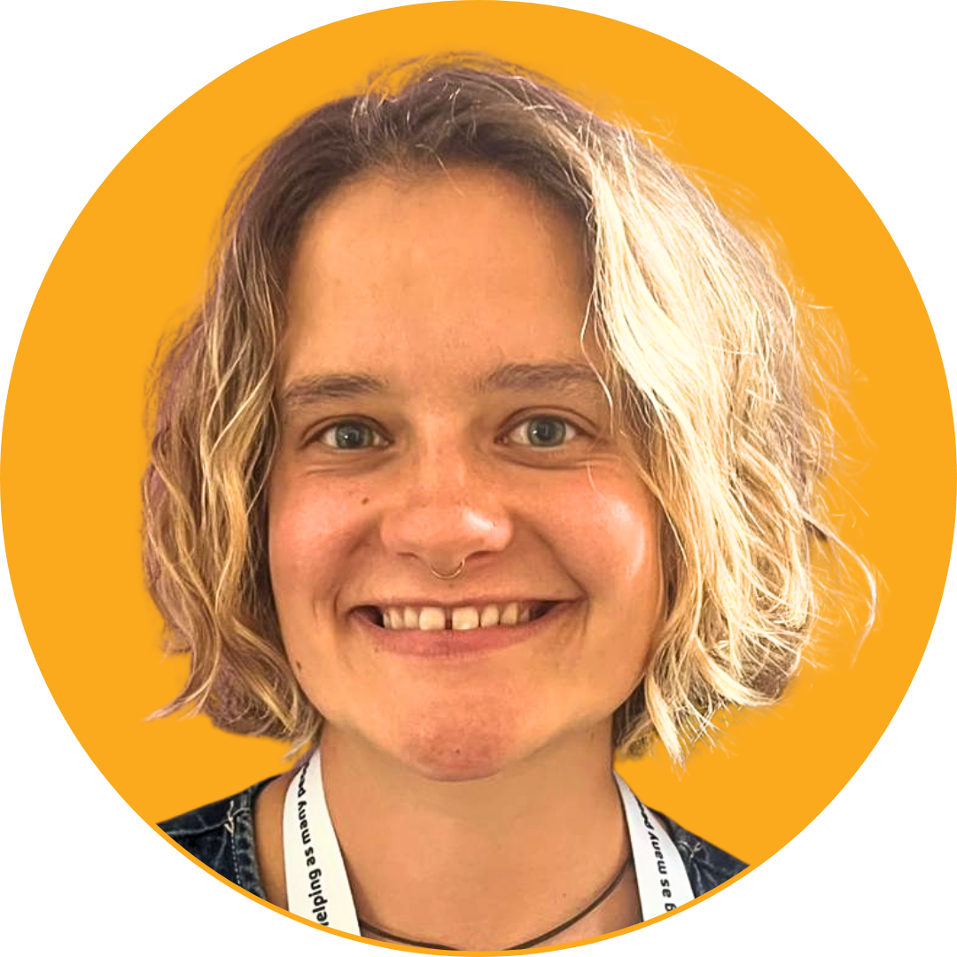 Close-up of a smiling young woman with short blonde curly hair, wearing a black top and a lanyard against an orange background.