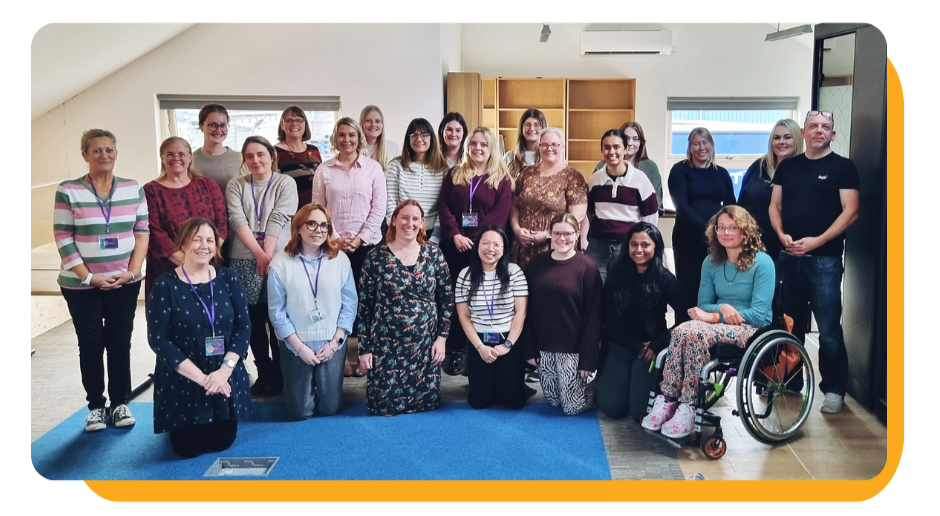 Group of diverse women and a man posing together indoors, some wearing lanyards, with one woman in a wheelchair, in a room with windows and wooden furniture.