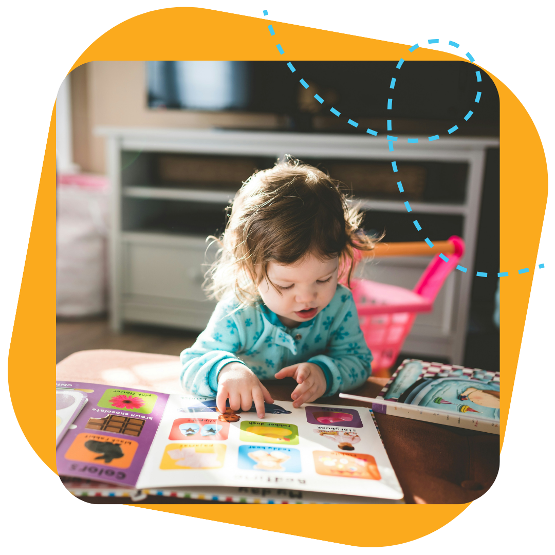 Young girl reading a colorful activity book at a table, with a pink shopping cart and TV stand in the background.