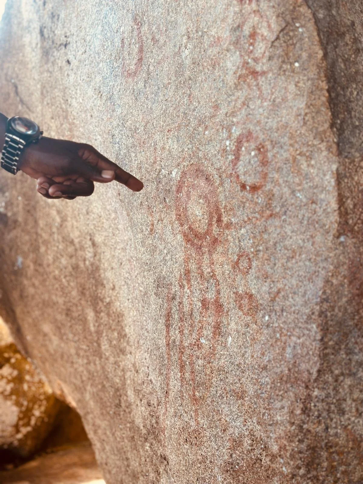 A person's hand with a watch points to a red painted symbol on a rough, brown, weathered rock surface.