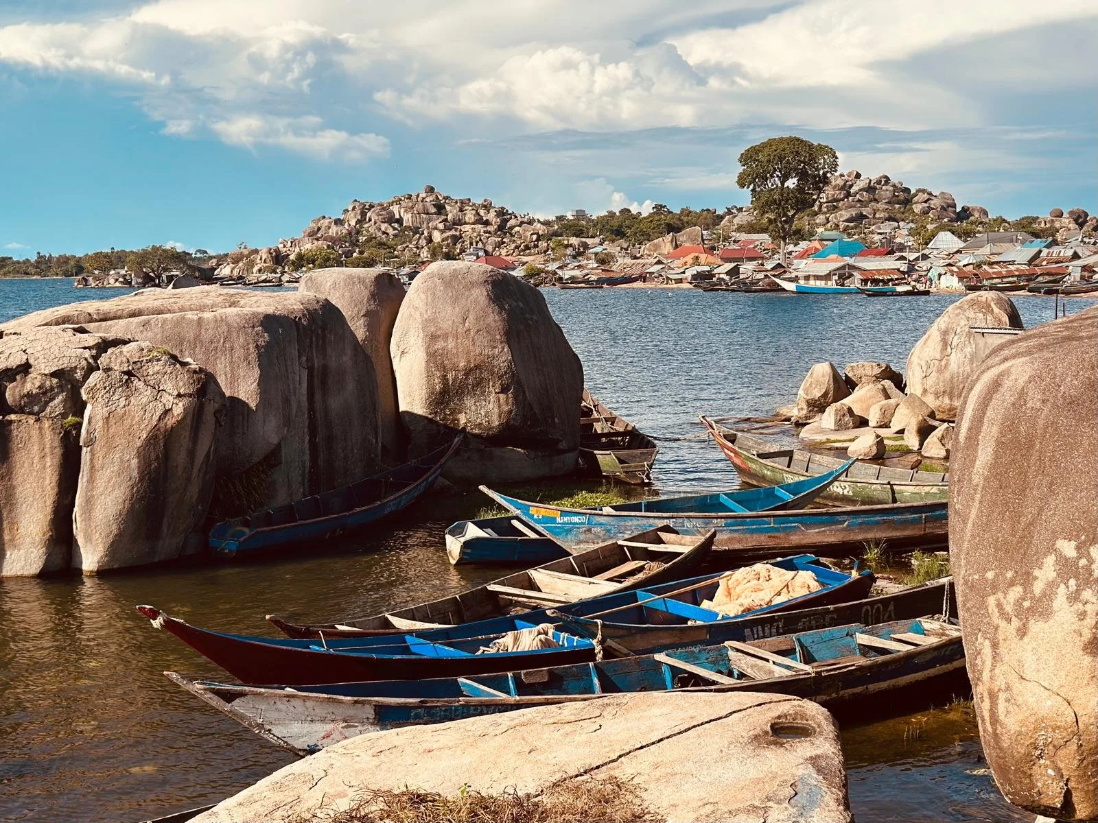 Boats docked along rocks on a lakeshore on Dolwe Island, with small houses and hills in the background under a partly cloudy sky.