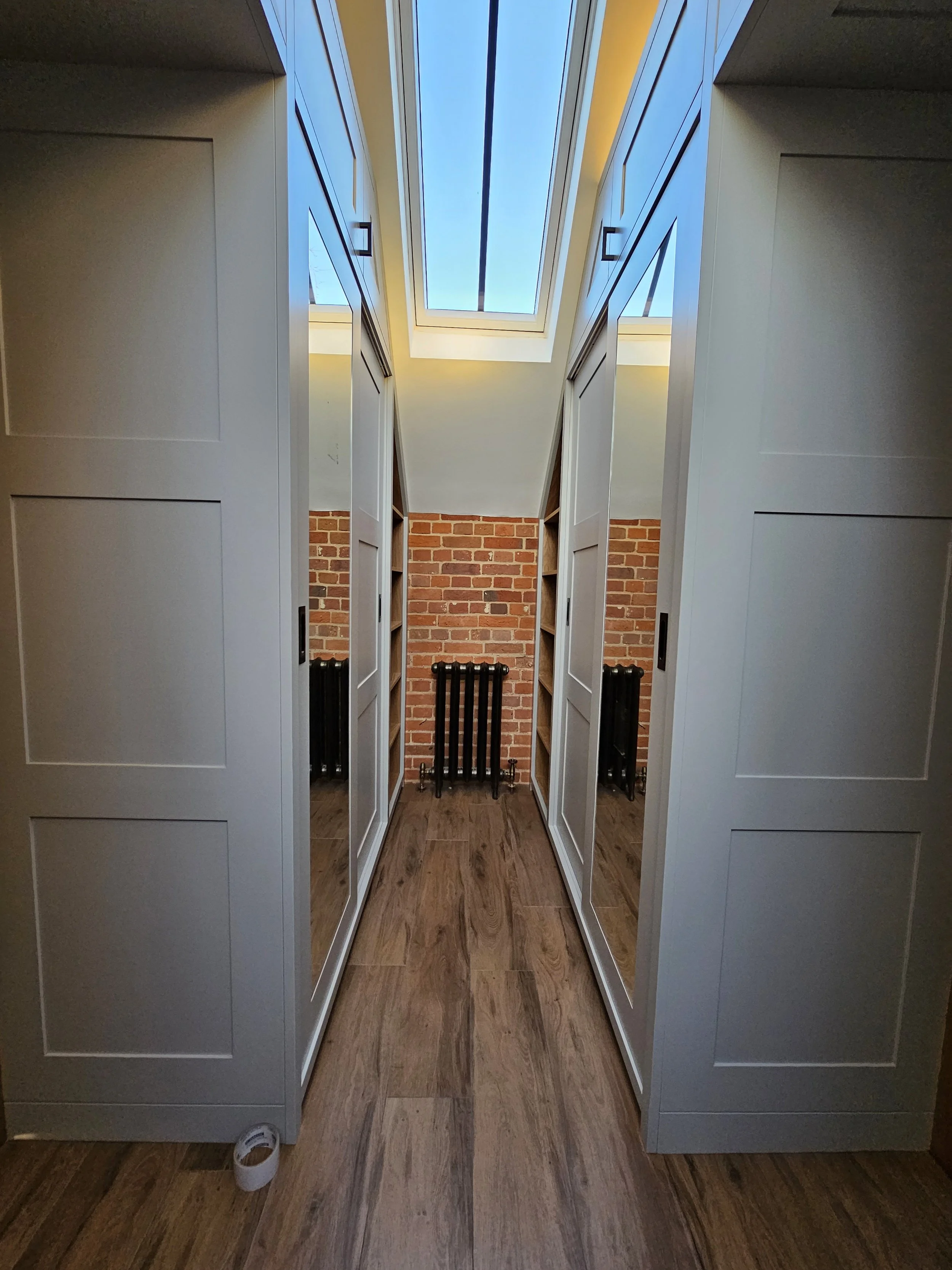 A narrow hallway with white cabinets on either side, a brick wall at the end, a skylight overhead, and a wooden floor.