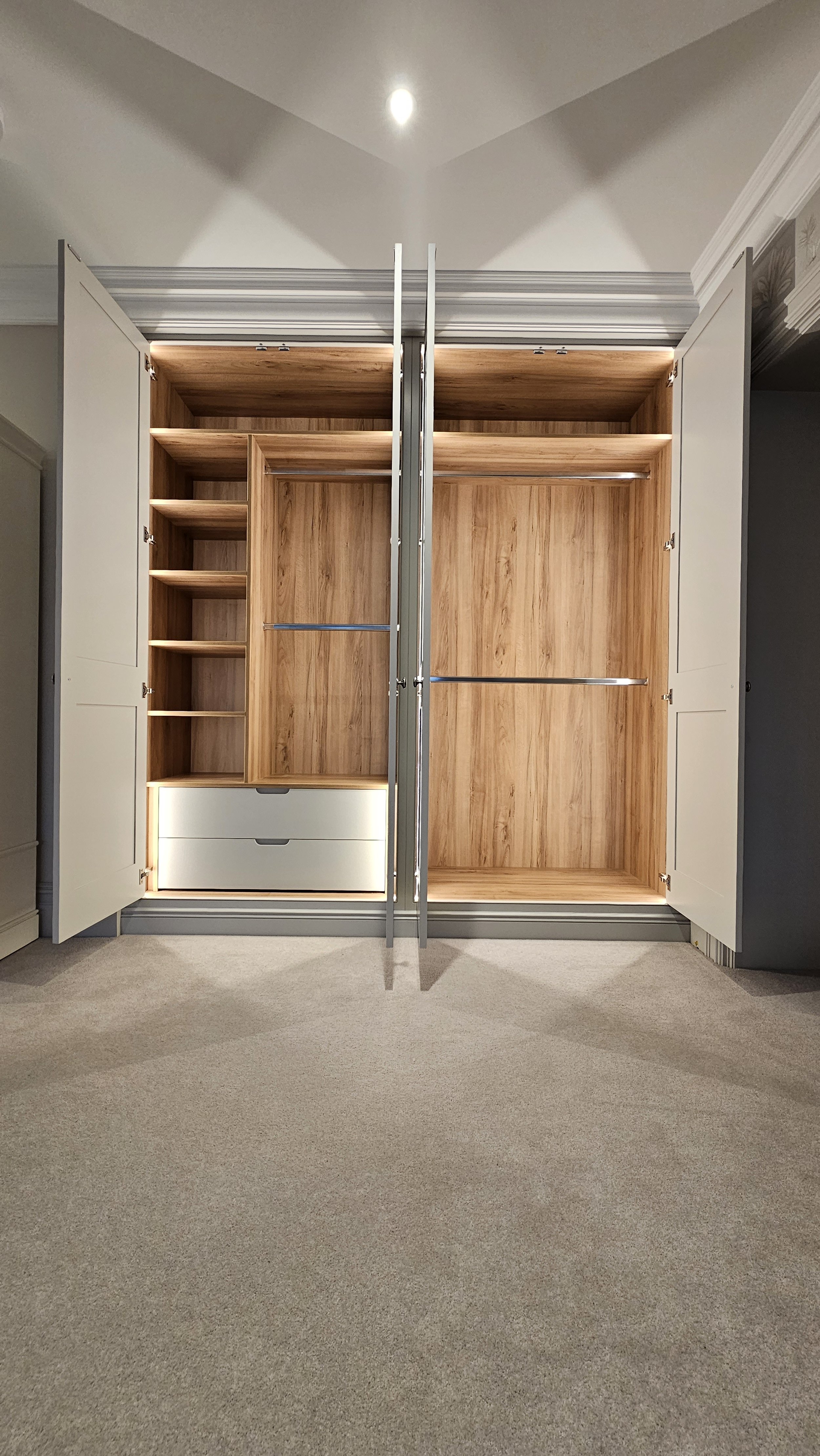 Open wardrobe with wooden interior, shelves, drawers, and hanging space, set against white doors and a beige carpeted floor.