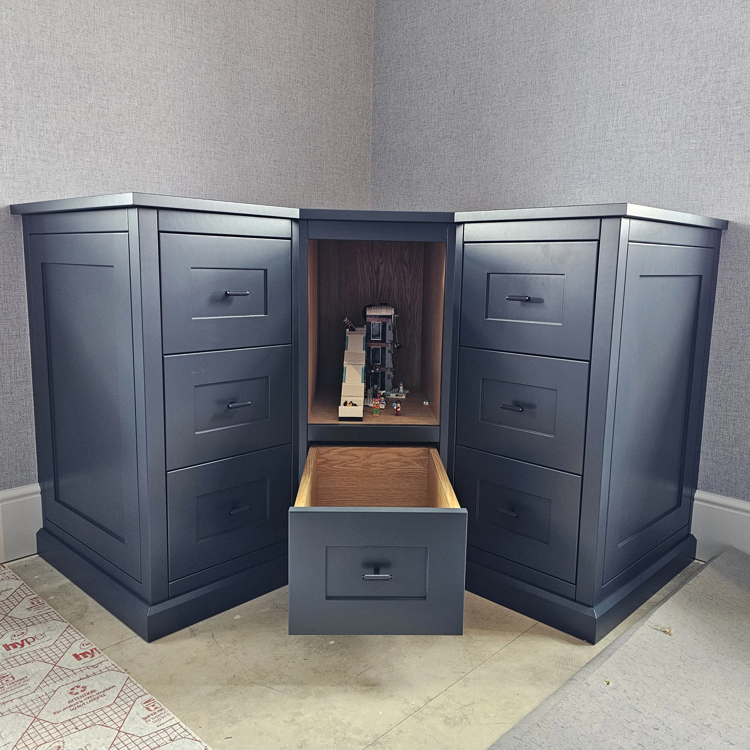 Dark blue wooden dresser with nine drawers and a central open compartment displaying LEGO sets, one drawer pulled out in front of it, against a gray wall.