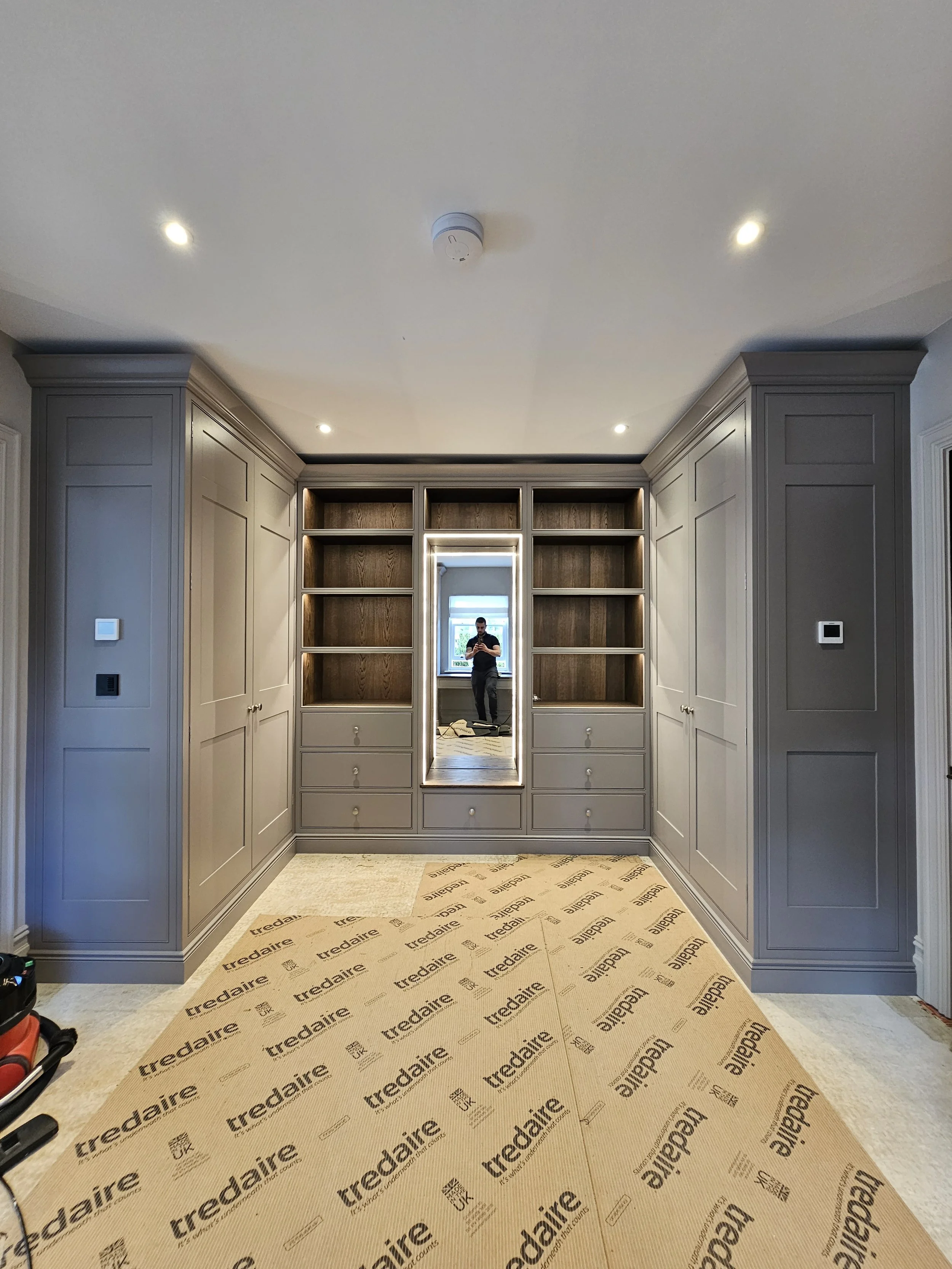 A hallway with built-in gray cabinetry on both sides, a mirror at the end reflecting a window and person, and a carpeted floor with a large cardboard sheet labeled 'Tredaire'.