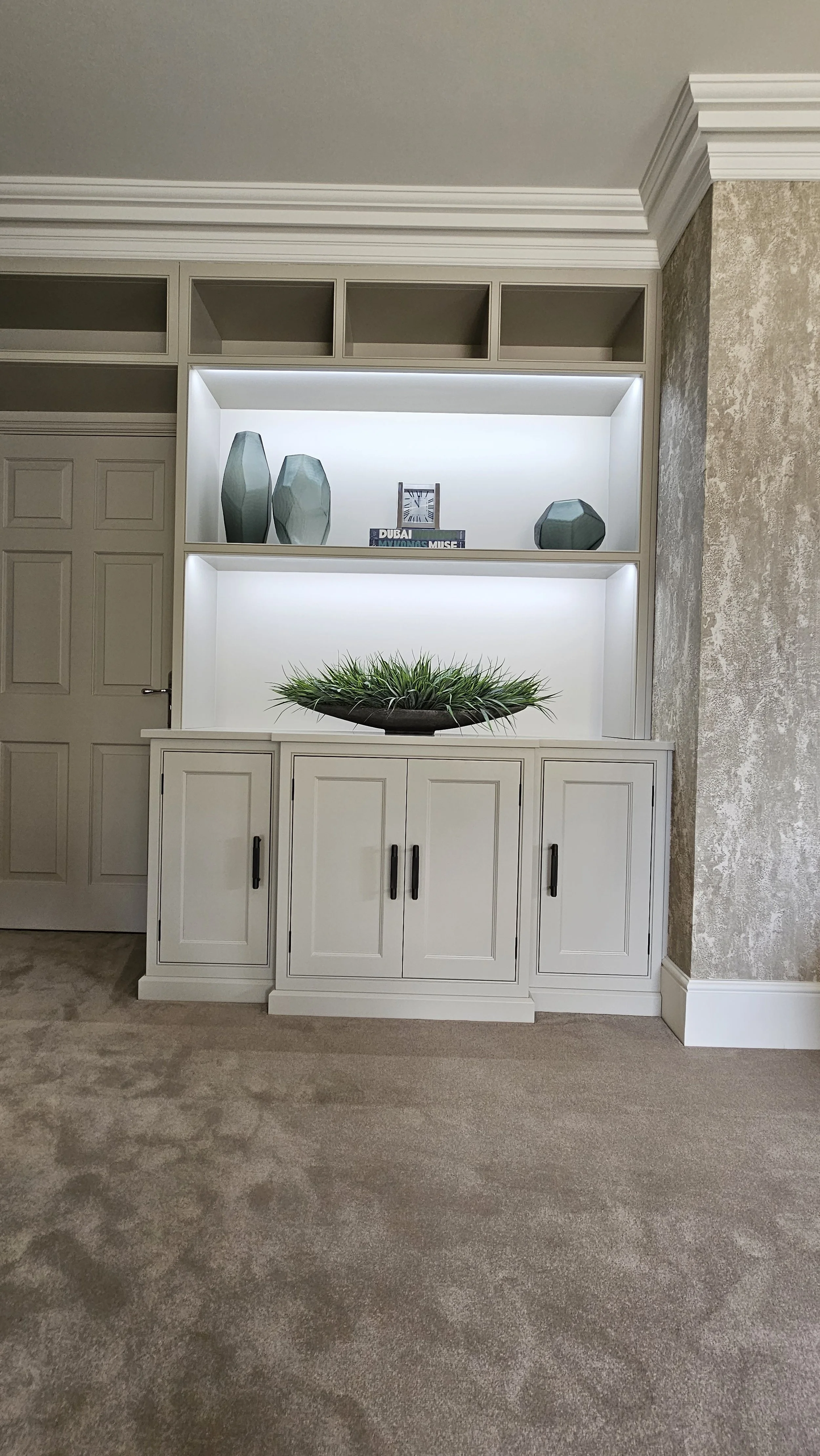 A built-in white cabinet with black handles, decorated with a large, shallow bowl of green ornamental grass, against a beige textured wall in a living room. The cabinet has open shelves above with decorative vases and books, illuminated with built-in
