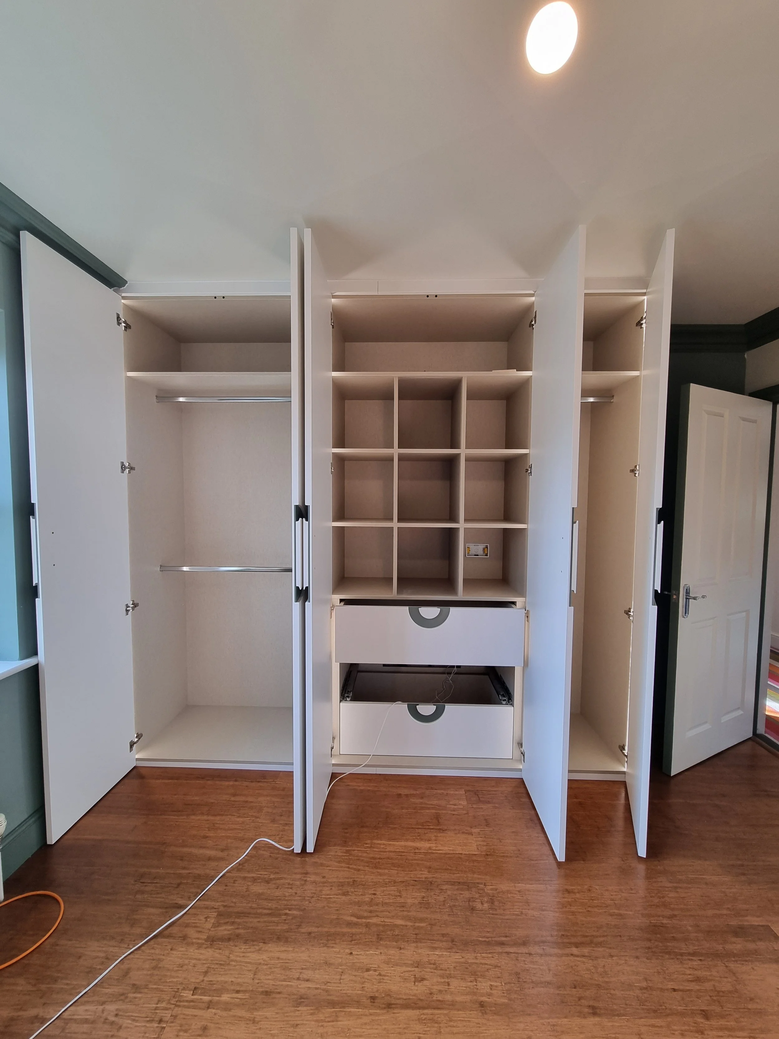 Empty white wardrobe with open doors, multiple sections, shelves, drawers, and hanging rods, set against a wall with a wooden floor.
