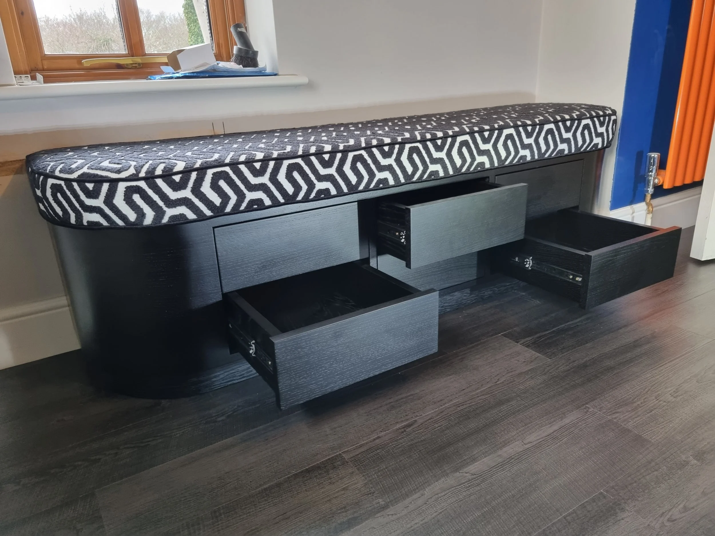 A black wooden console table with three open drawers and a padded black and white patterned top against a wall with a window and a blue wall on the right.