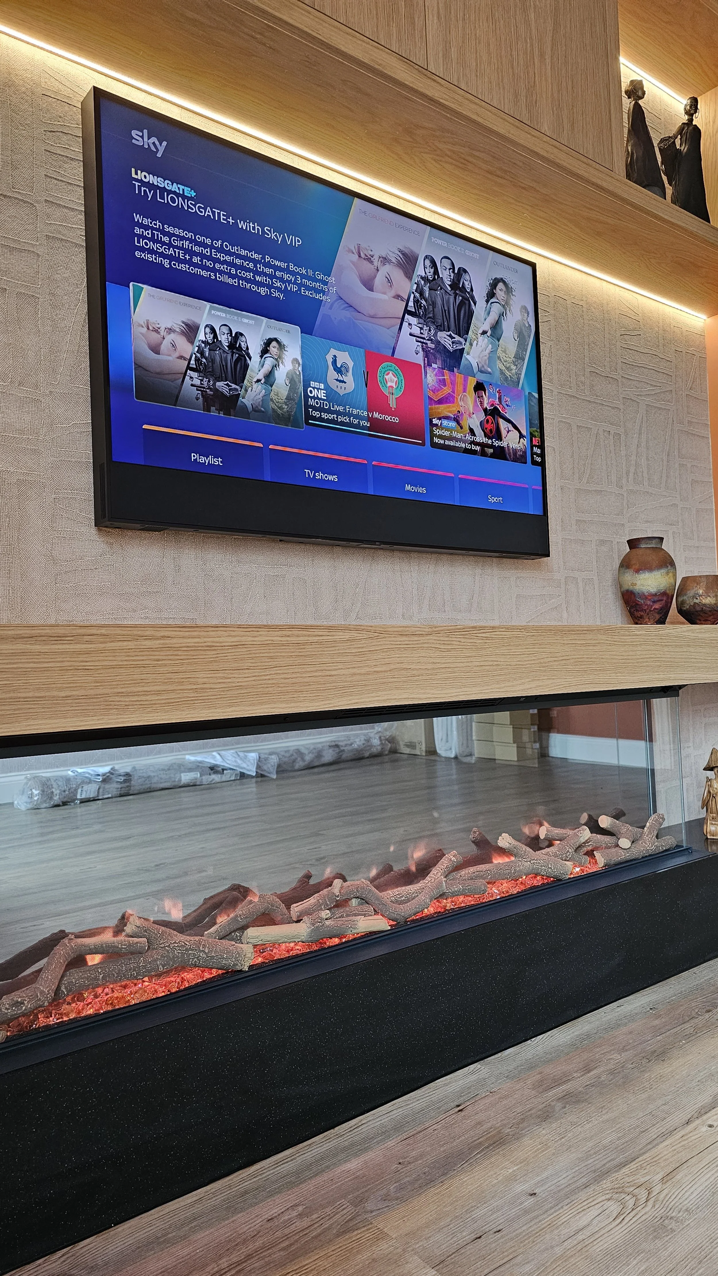 A living room with a flat-screen TV mounted on the wall showing a streaming service menu, and a modern electric fireplace below it with decorative logs. There are two small vases on a wooden shelf above the fireplace.