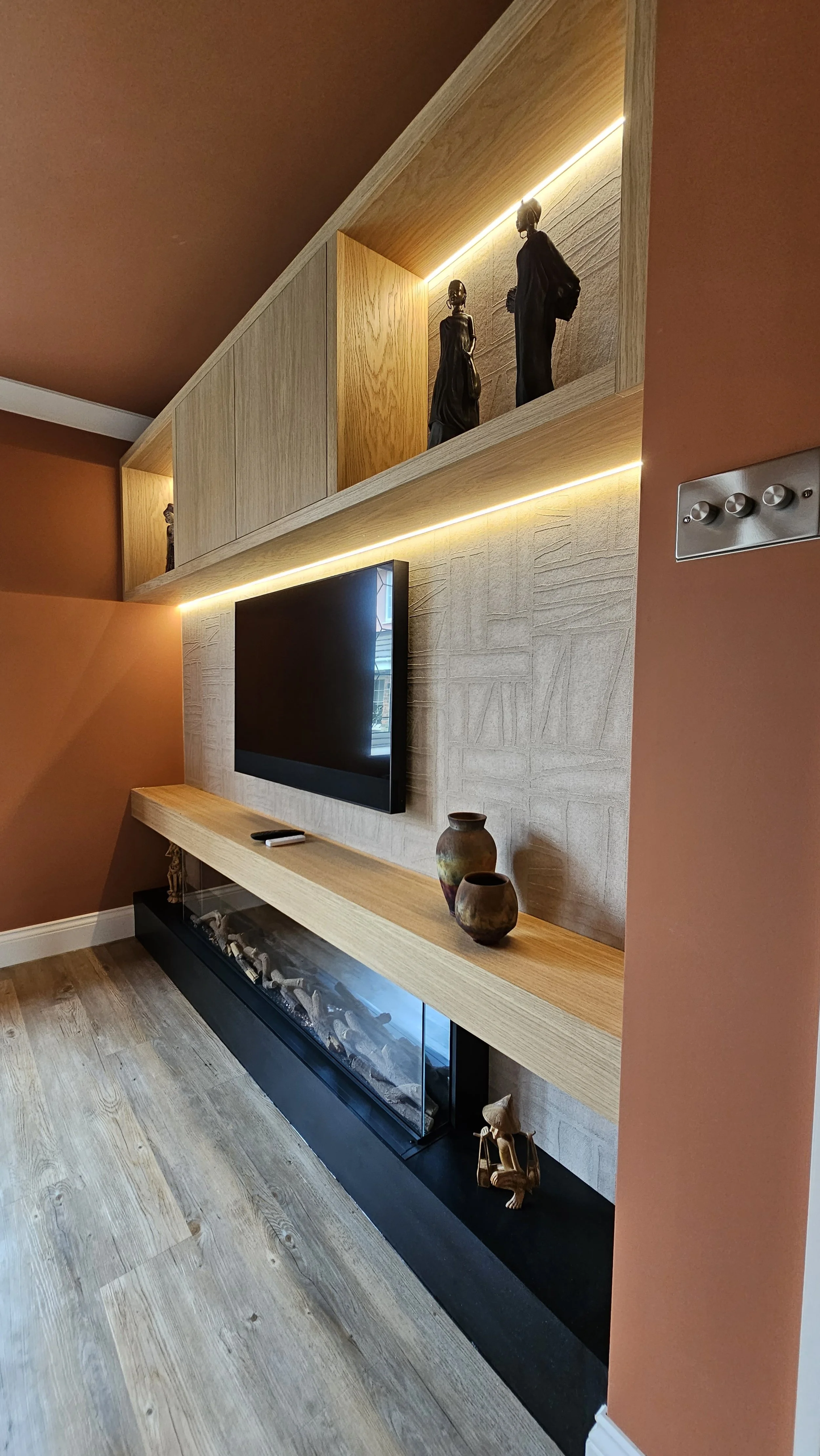Living room wall with a mounted TV, wooden shelves and decorative vases, and a fireplace below with logs inside.