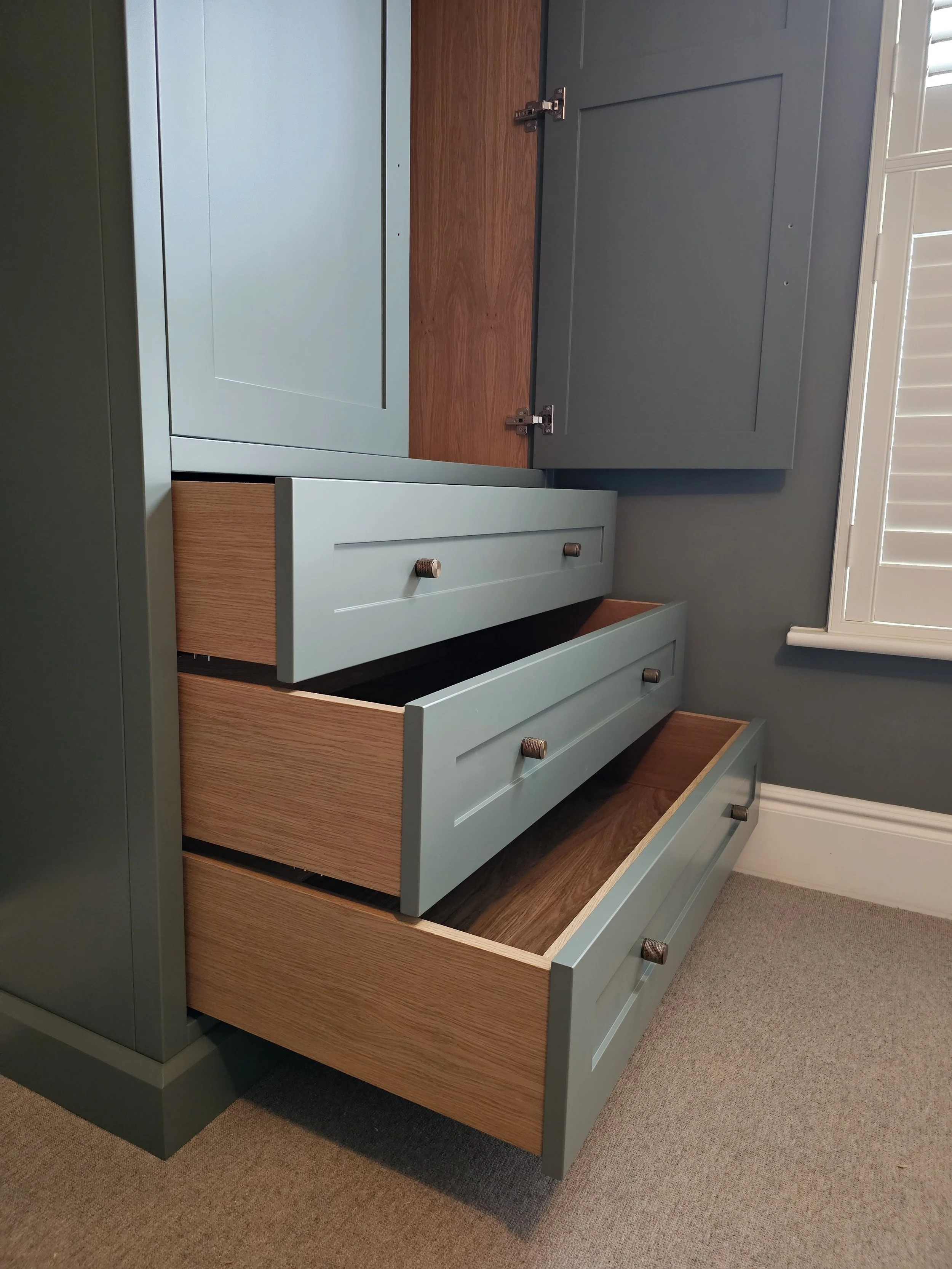 Open drawers of a modern dresser with light blue fronts and wooden interiors, situated next to a gray wall and window with white shutters.
