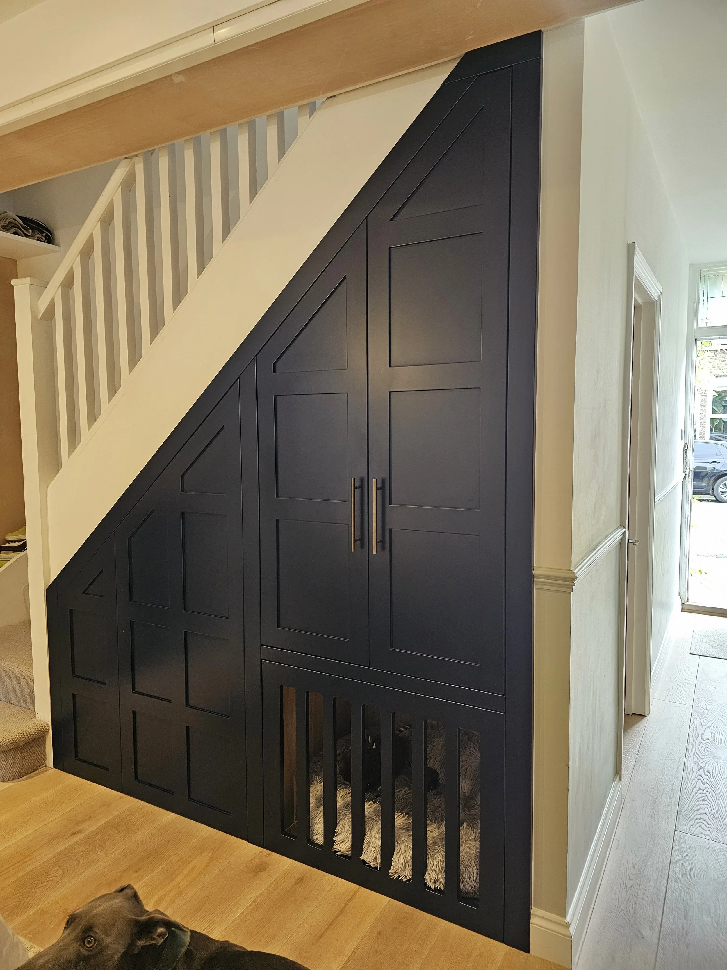Built-in black storage cabinet under a staircase with a dog resting underneath, in a home's interior hallway.