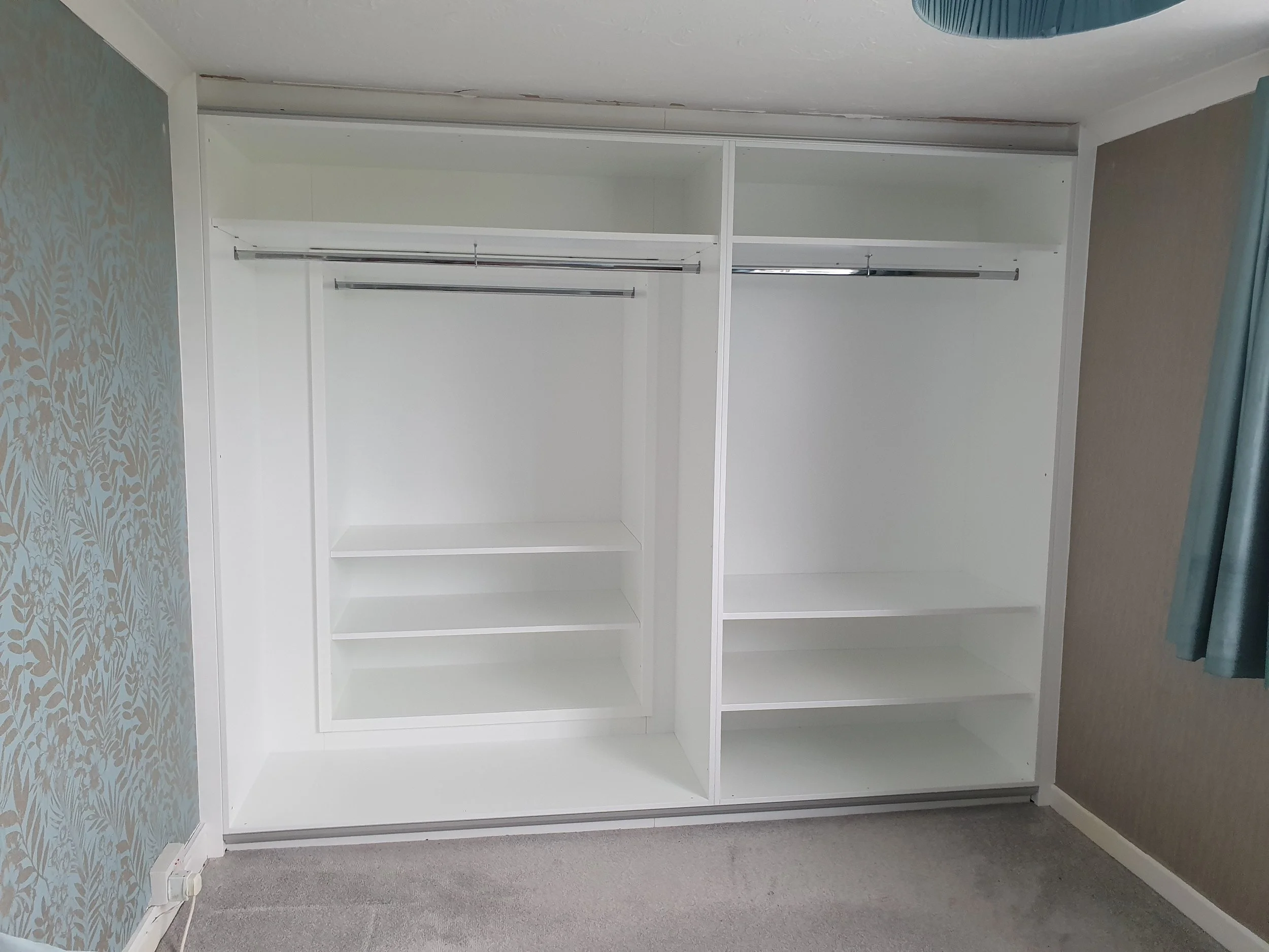 Empty white built-in closet with shelves and hanging rods, in a room with patterned and plain wallpaper, blue curtains, and gray carpeted floor.