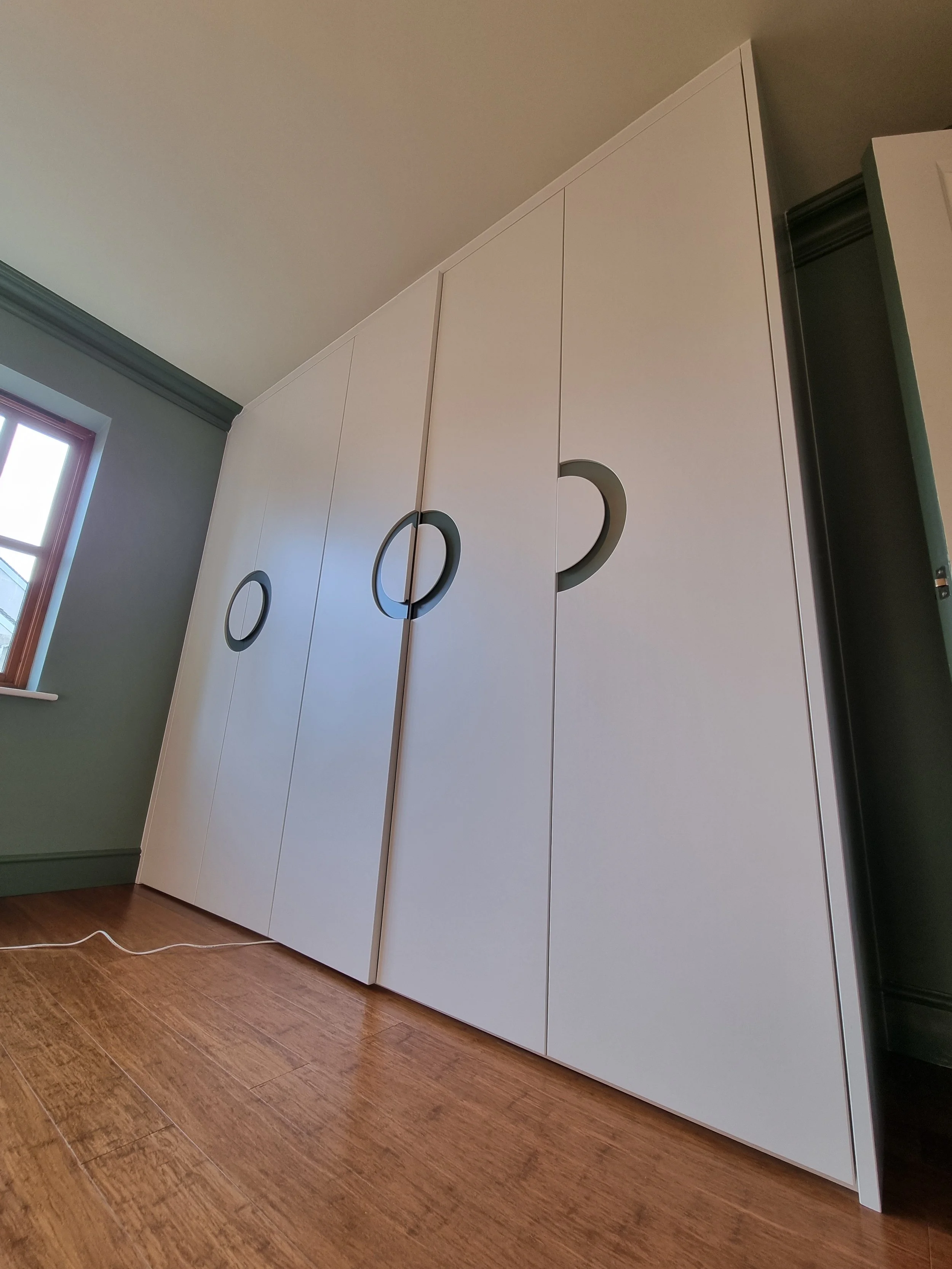 White wardrobe with circular cutout handles in a room with hardwood floor and a window.
