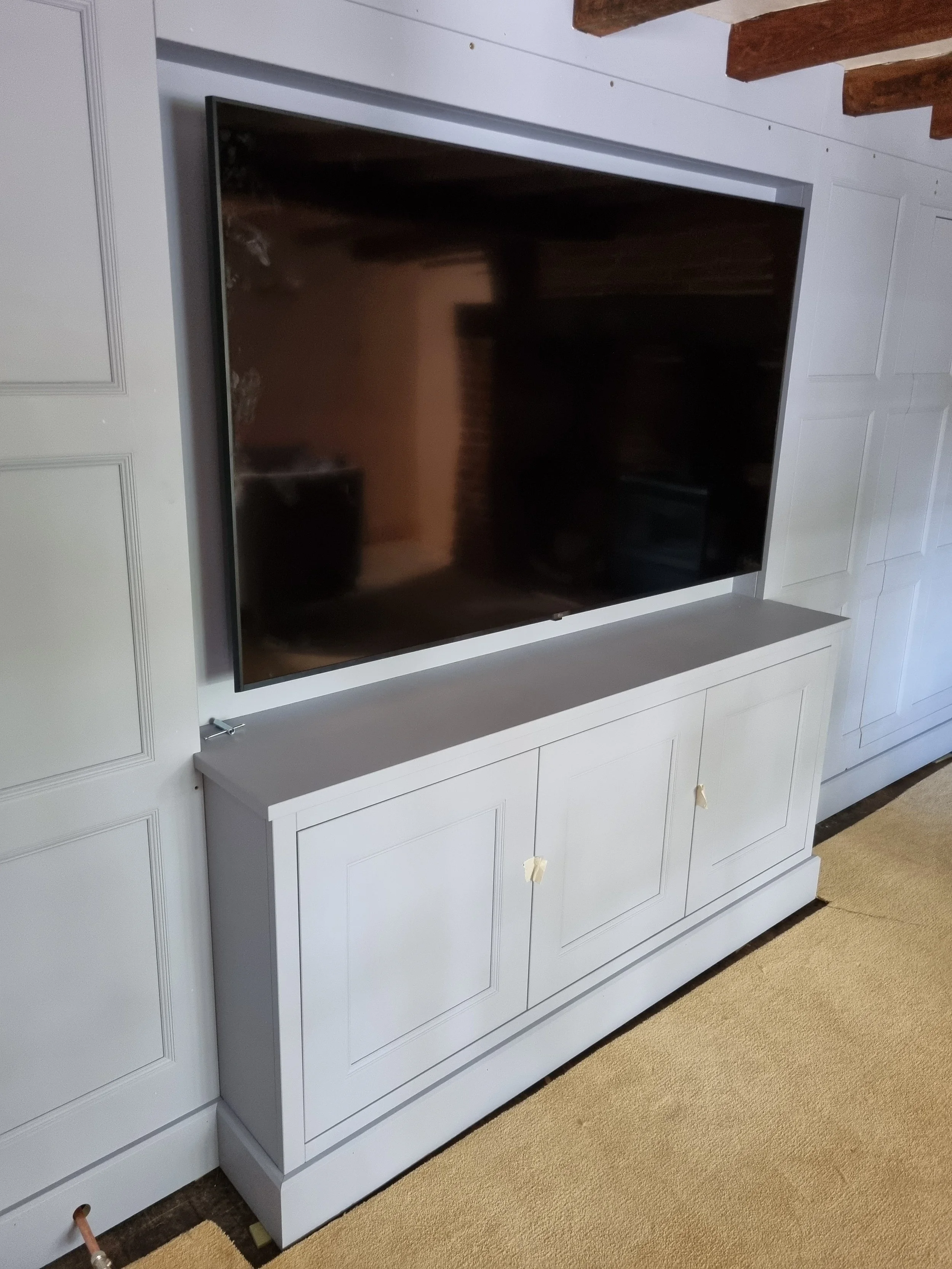 A large flat-screen TV mounted on a white cabinet with double doors. The TV is turned off and reflects part of the room. The cabinet is against a white wall with decorative panels. There are wooden beams on the ceiling and a beige carpet on the floor