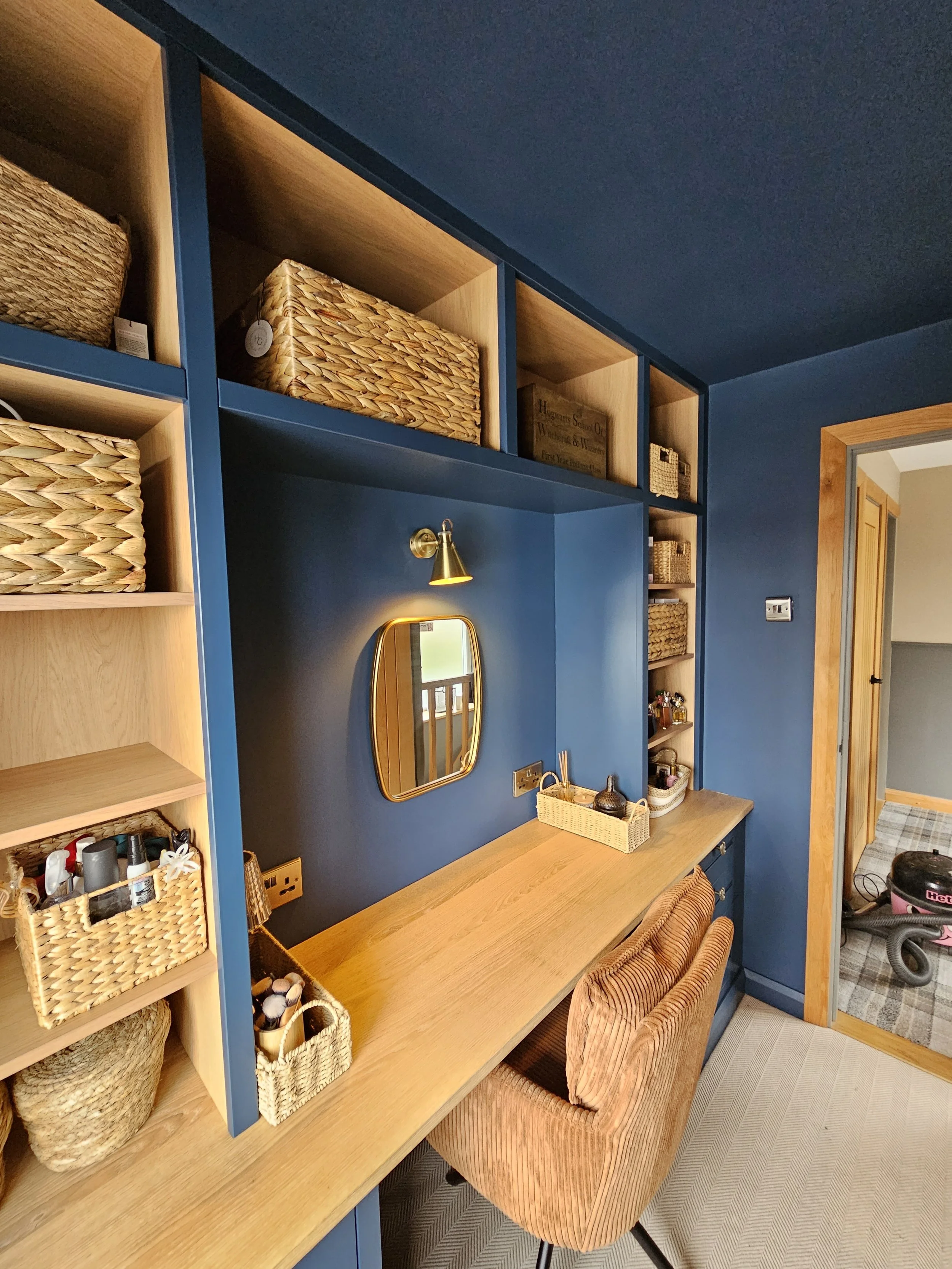 A cozy home office corner with a wooden desk, a peach-colored velvet chair, a small mirror, and blue wall with open shelves filled with baskets and decor.