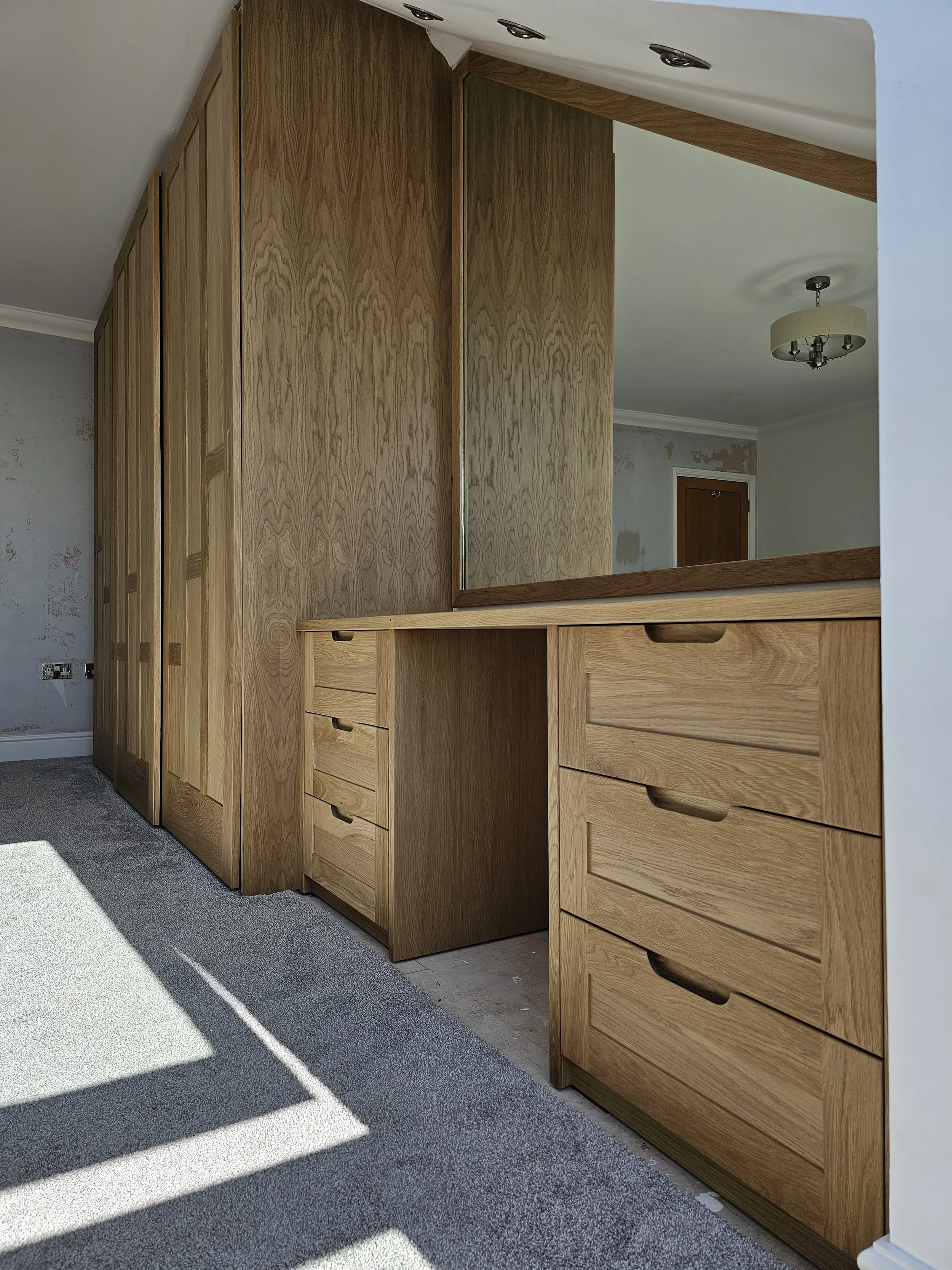 Wooden built-in wardrobe and dresser unit in a room with grey carpeting and partially painted wall.