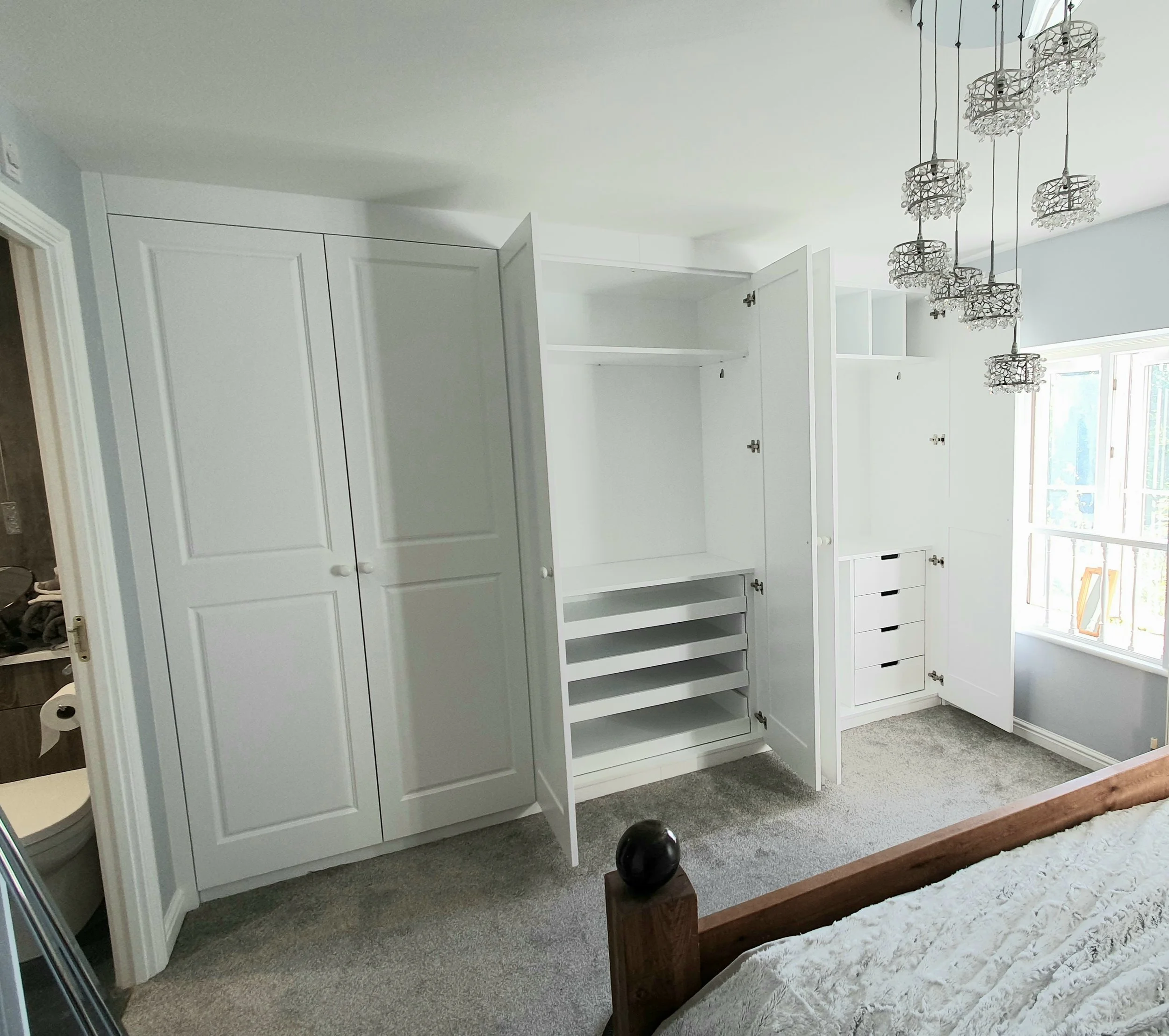 Empty built-in white closet with open doors, multiple shelves, and drawers in a bedroom next to a window with sunlight.