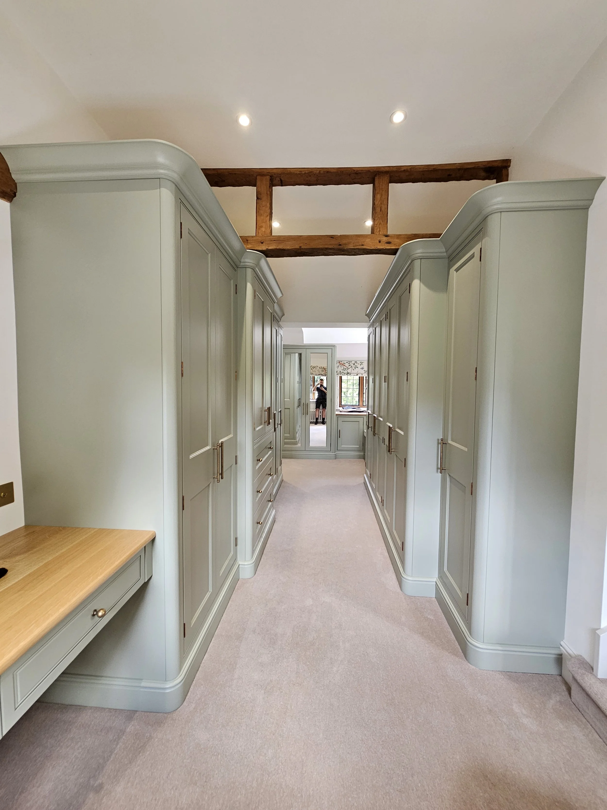 A walk-in closet with pale green built-in cabinets, carpeted floor, and wooden beams on the ceiling, leading to a mirror and window at the end of the room.