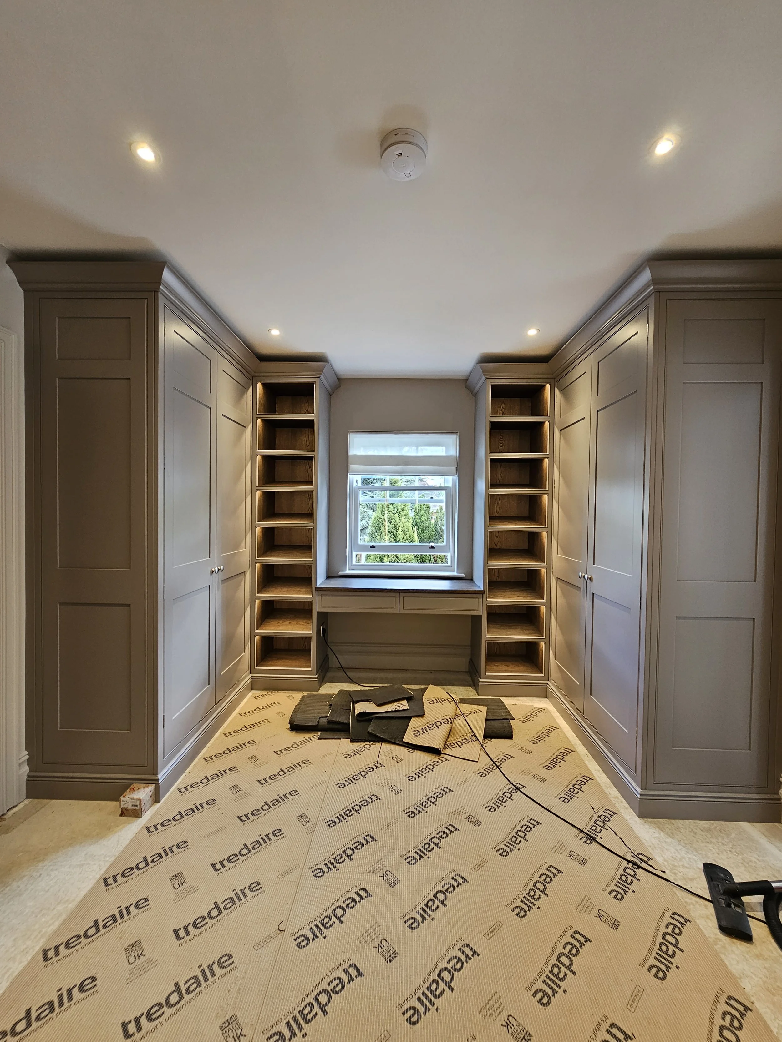 A room with built-in gray cabinetry, large open shelves, a window, and a workspace area, with flooring under construction.
