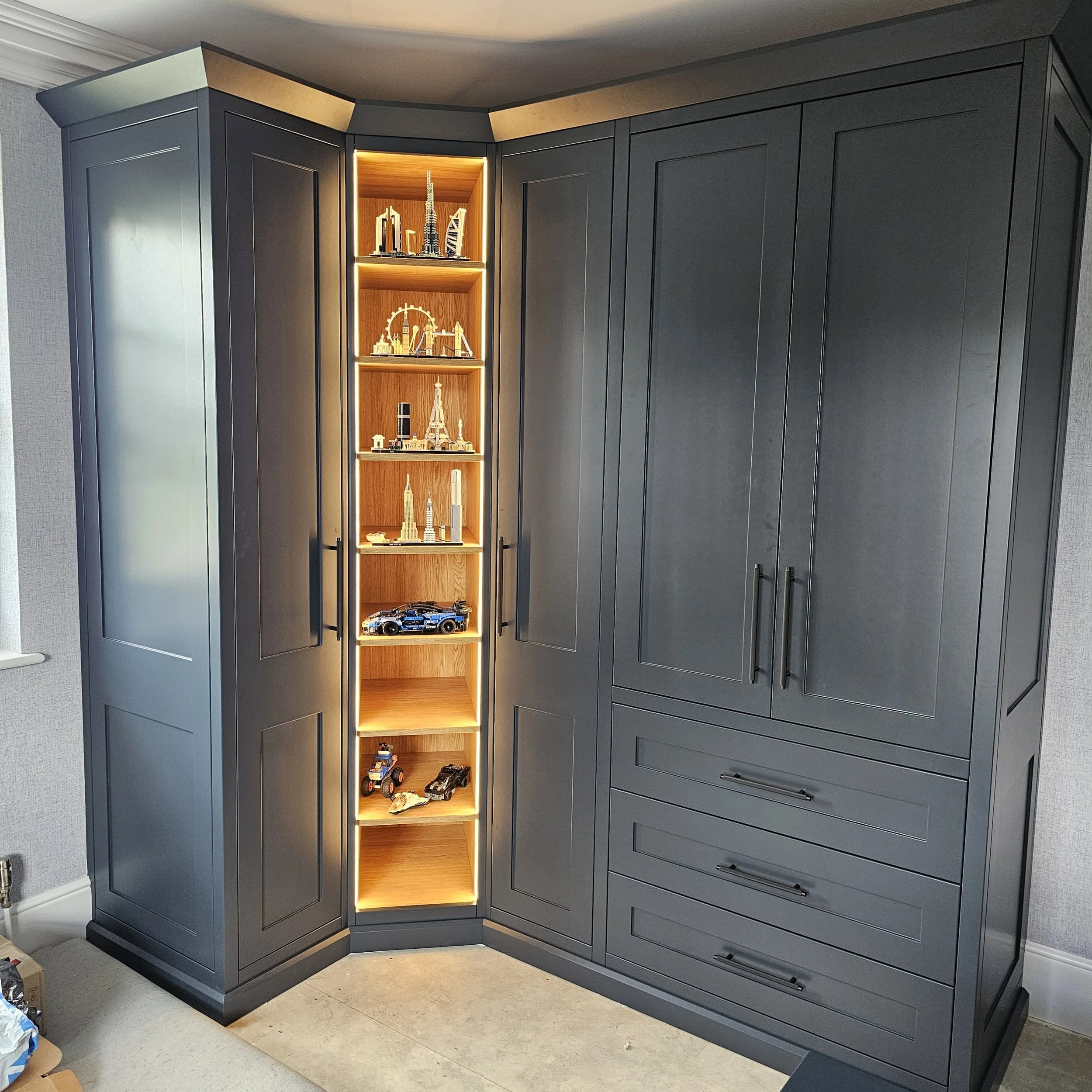 A corner of a room features a dark gray cabinet with multiple drawers and doors, next to a built-in illuminated display shelf with a wood interior containing miniature models of famous landmarks and toy cars.