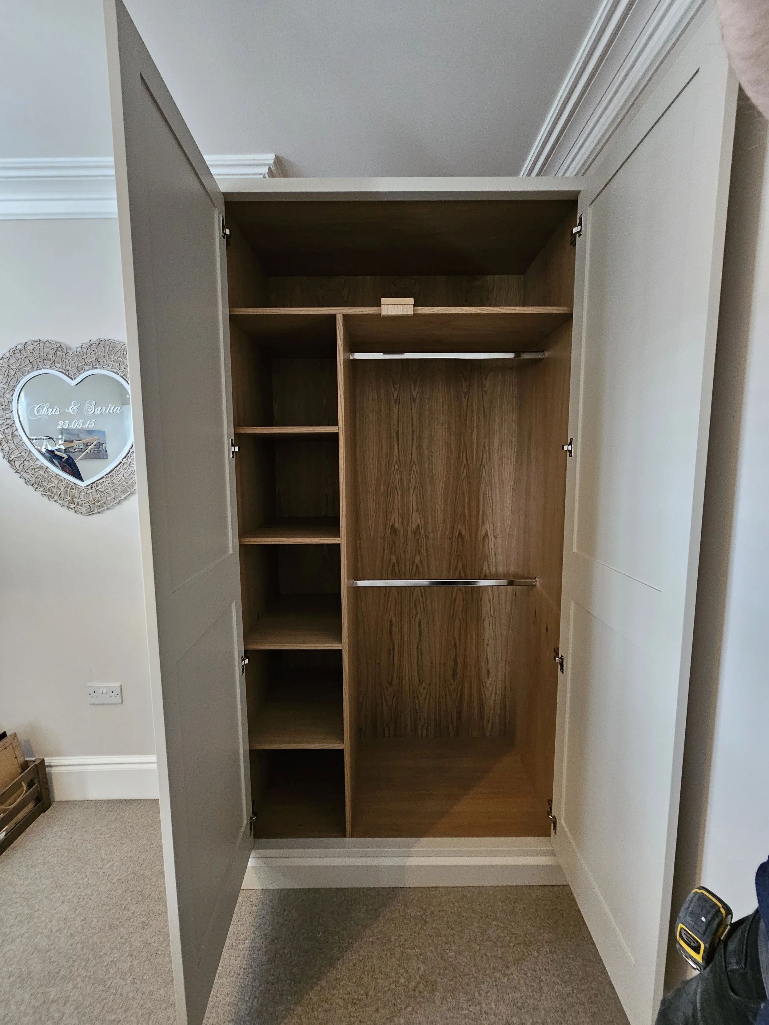 Open wardrobe with multiple shelves and hanging rods in a room with carpeted floor and a decorative wall mirror.