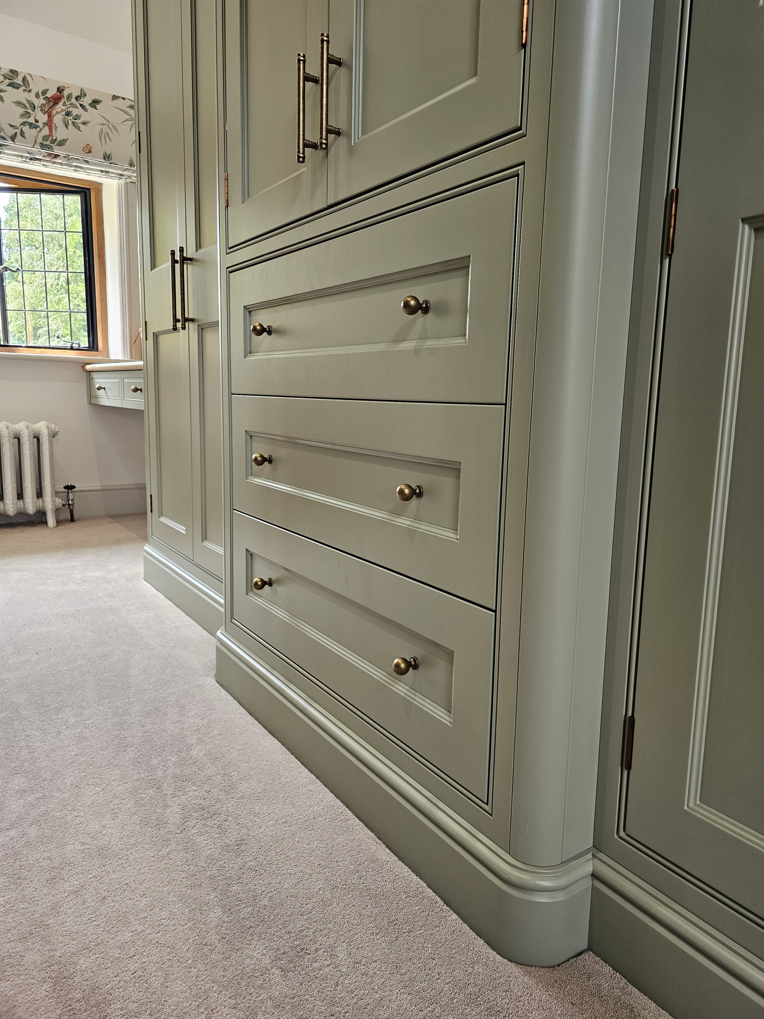 Close-up of pale green built-in cabinetry with gold knobs in a room with beige carpet, a window, and floral wallpaper.