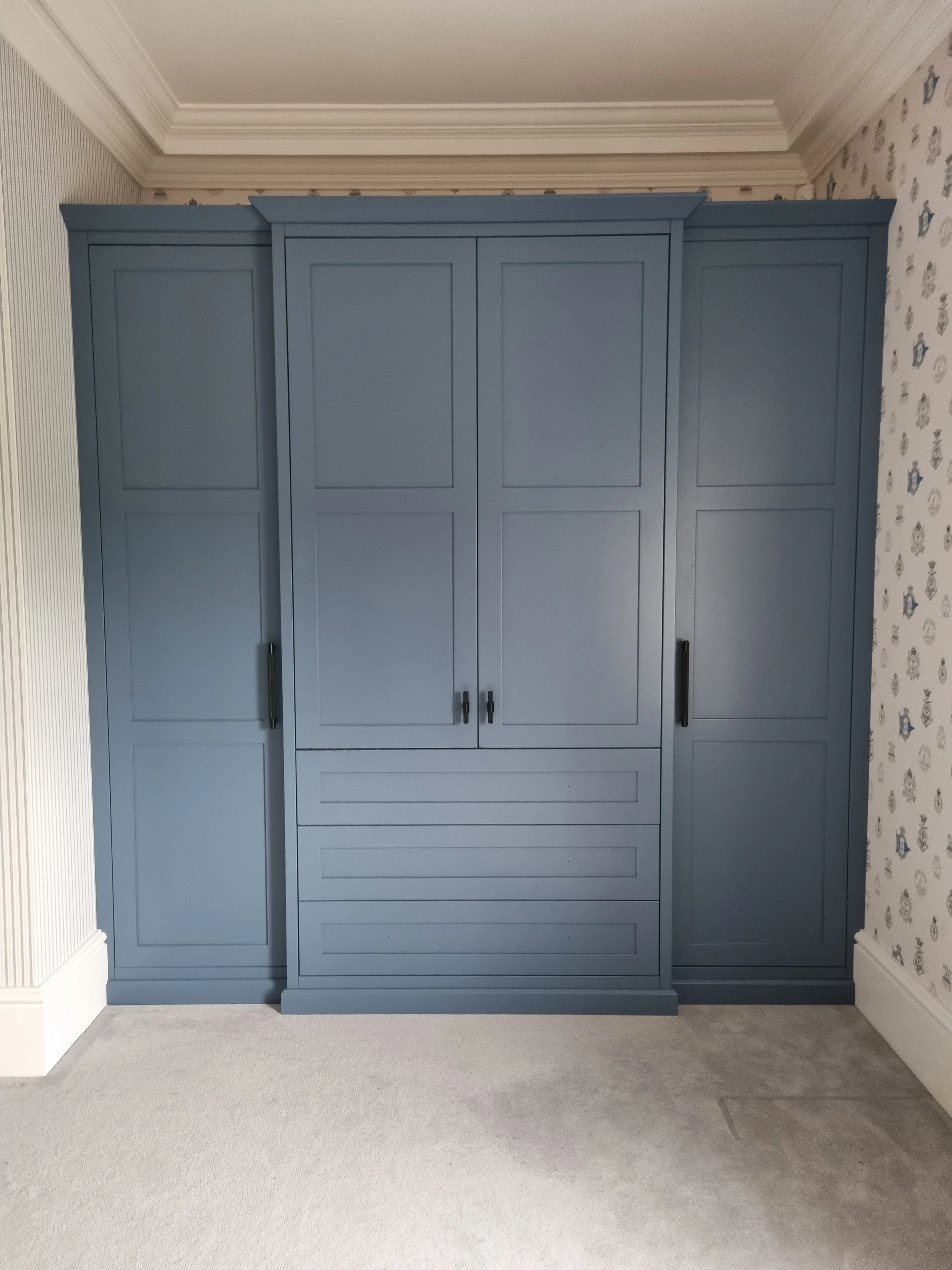 A large blue wardrobe with four doors, two drawers at the bottom, and black handles, placed in a room with patterned wallpaper and white crown molding.