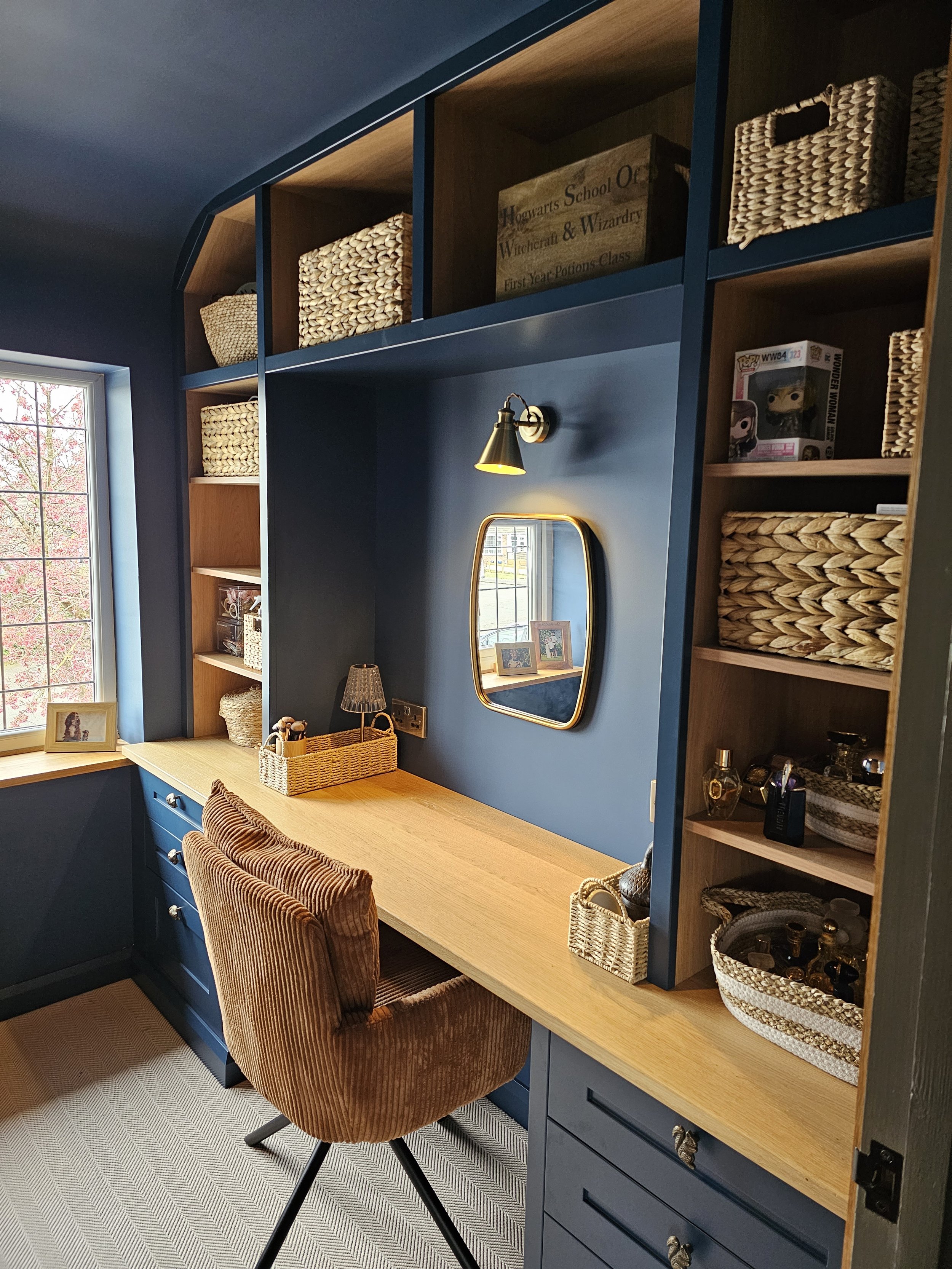 Home office or study area with blue walls, a wooden desk, a rust-colored upholstered chair, a window with a view of trees, and open shelves with woven baskets and decorative items.