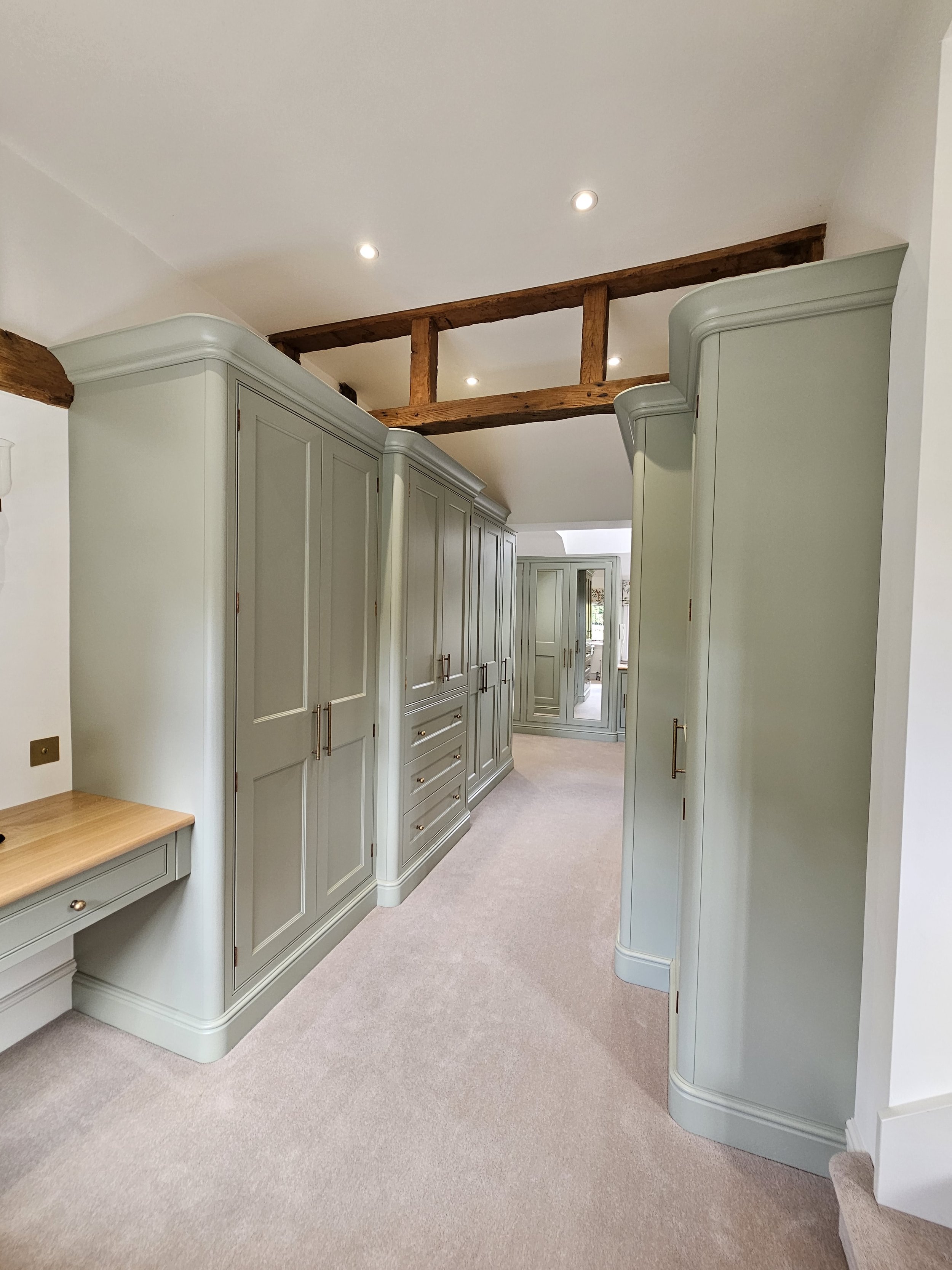 A hallway lined with tall, pastel green built-in cabinets with gold handles, beige carpet, and wooden ceiling beams, leading to another room with similar cabinets.