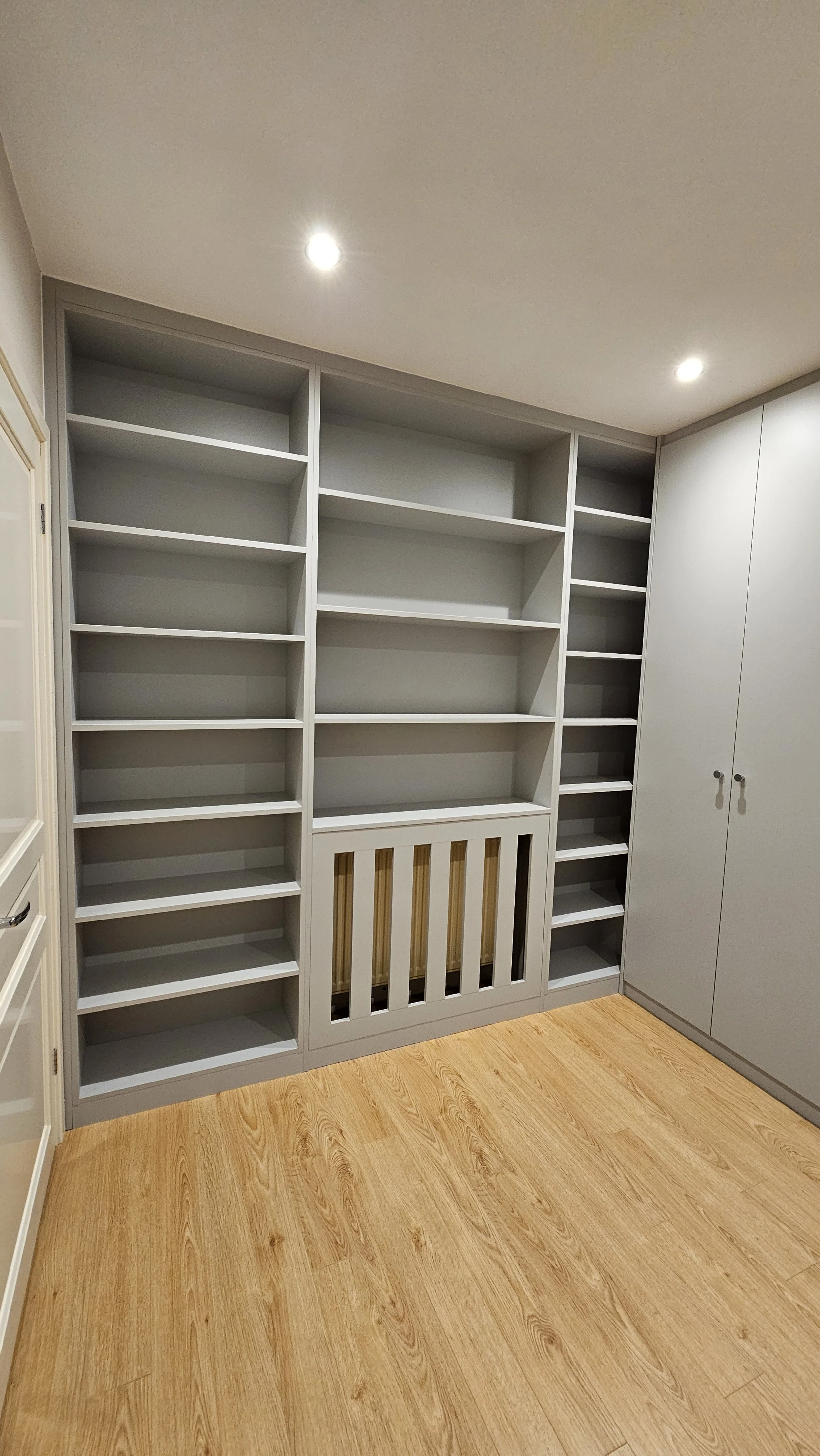 Built-in white shelving unit with open shelves and a small slatted door, next to a white closet with double doors, in a room with light wood flooring and ceiling spotlights.