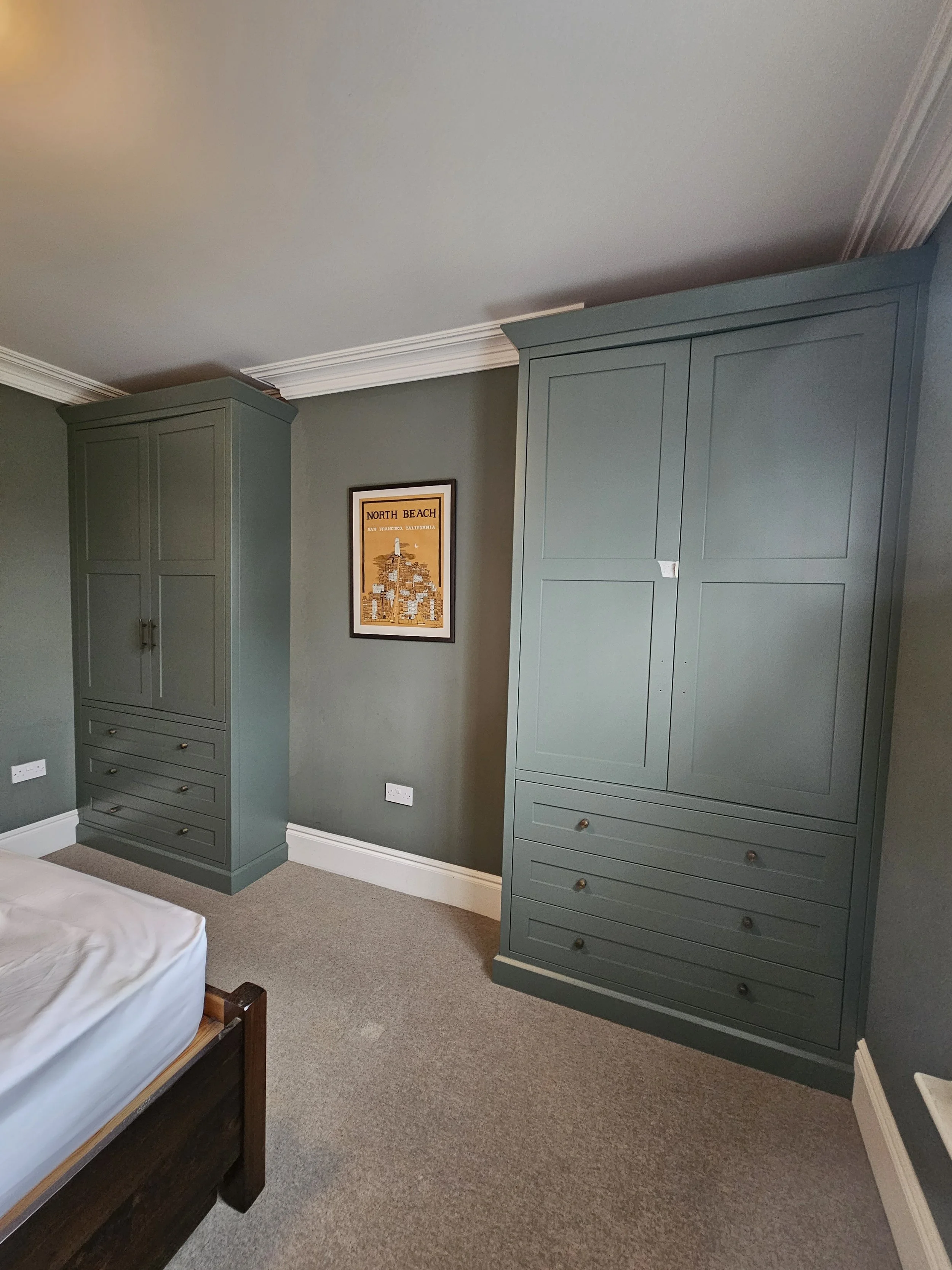 A bedroom corner with two large green wardrobes, a framed North Beach art print on the wall, a corner of a bed with a white mattress, beige carpet, and white crown molding.