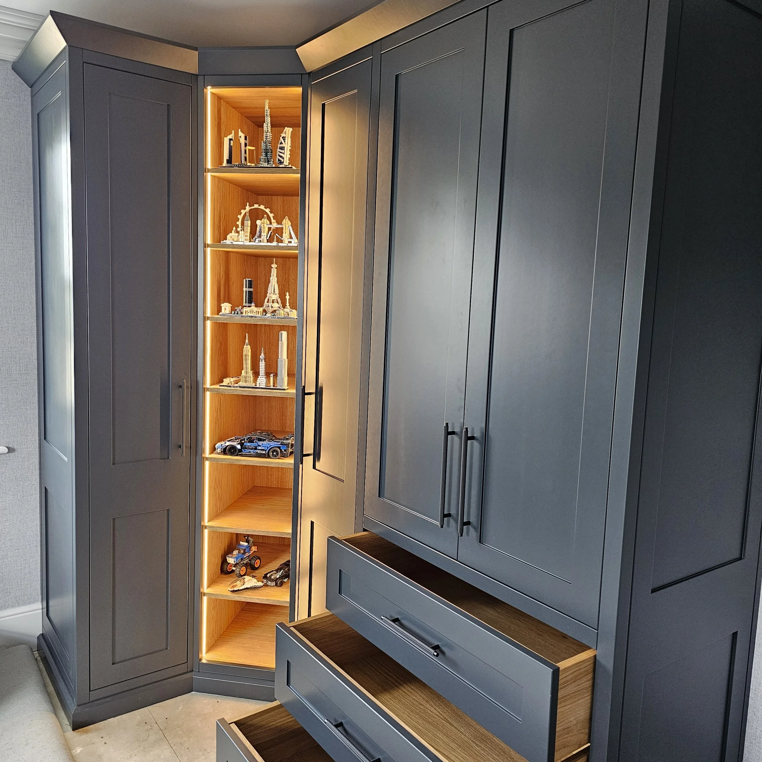 A dark gray corner wardrobe with paneled doors and black handles. One section has open wooden shelves displaying miniature models of London landmarks and cars, with warm interior lighting.