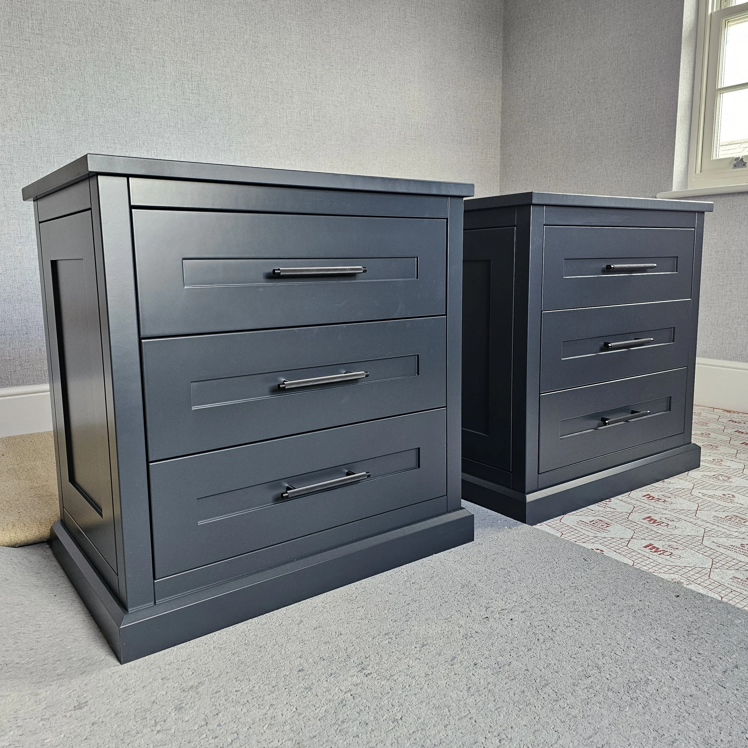 Two dark gray bedside tables with three drawers each, placed on a beige carpet in a room with light gray walls and a window in the background.