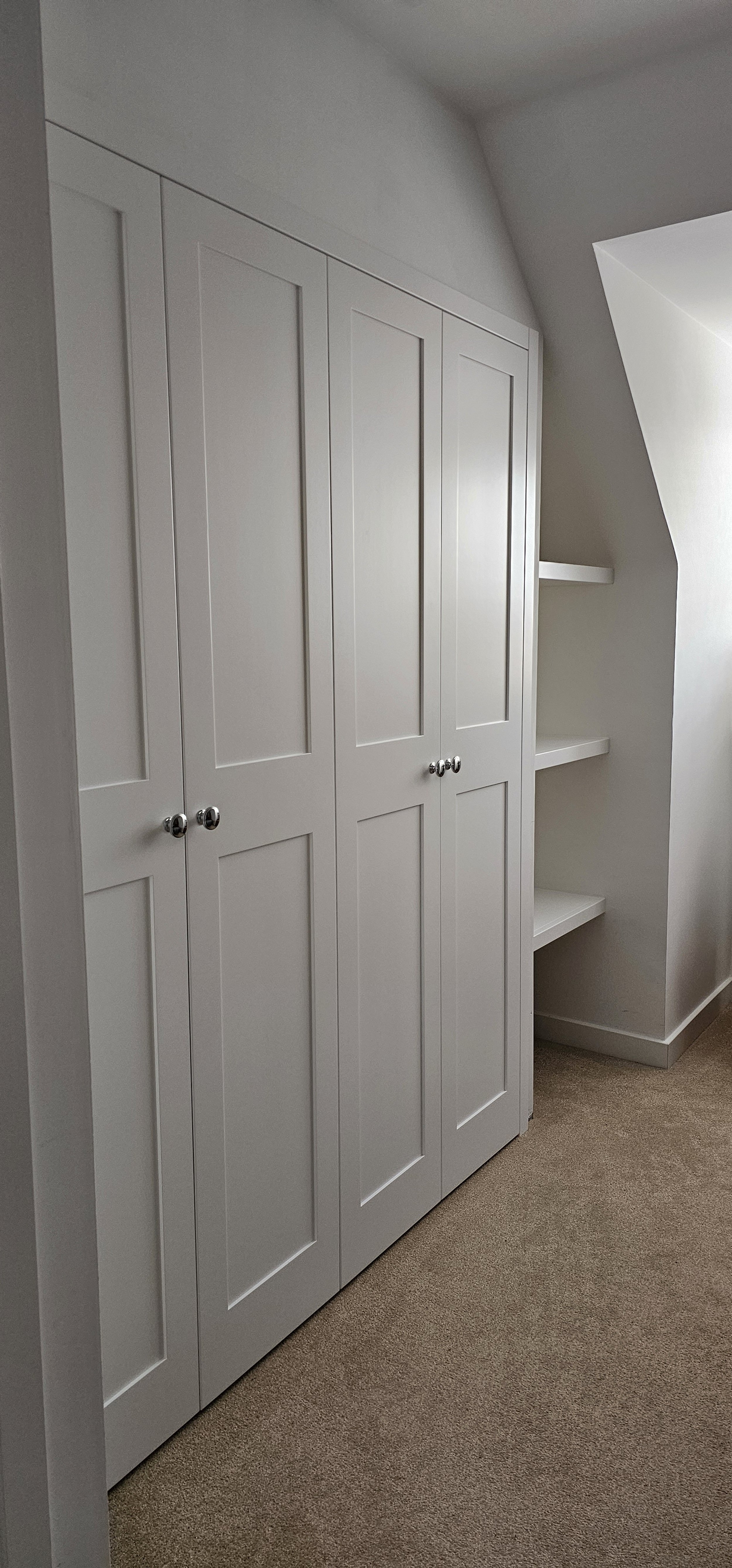 White built-in closet with four panels and round metallic knobs, adjacent to white floating shelves on the wall, in a room with carpeted floor.