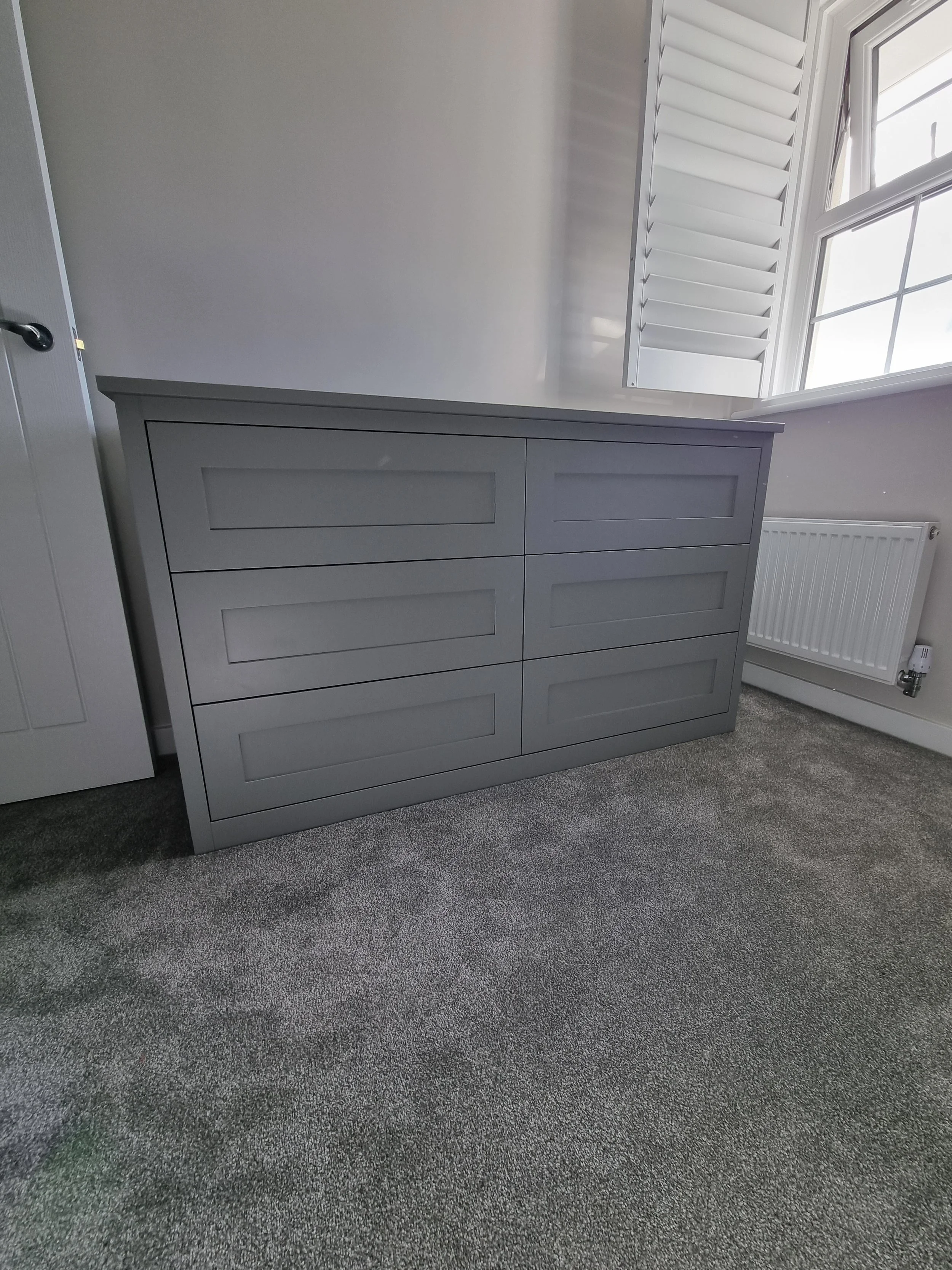 Gray dresser with six drawers in a room with gray carpet and a window with white shutters.